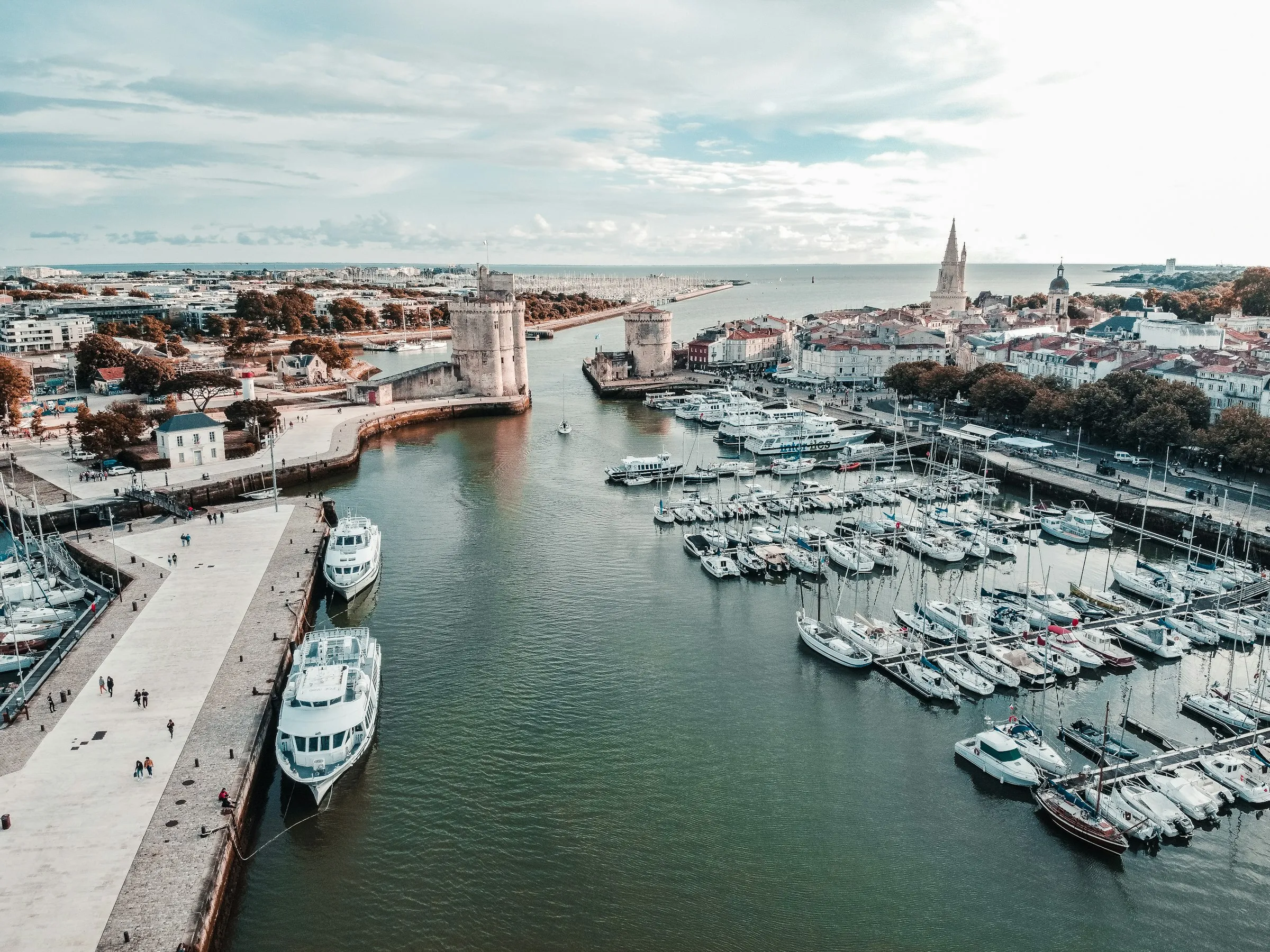 Aerial view of La Rochelle's medieval harbour mouth guarded by the Tour Saint-Nicolas and Tour de la Chaine, with the Tour de la Lanterne spire, a marina packed with white yachts and the terracotta-roofed old town spreading to the Atlantic beyond.