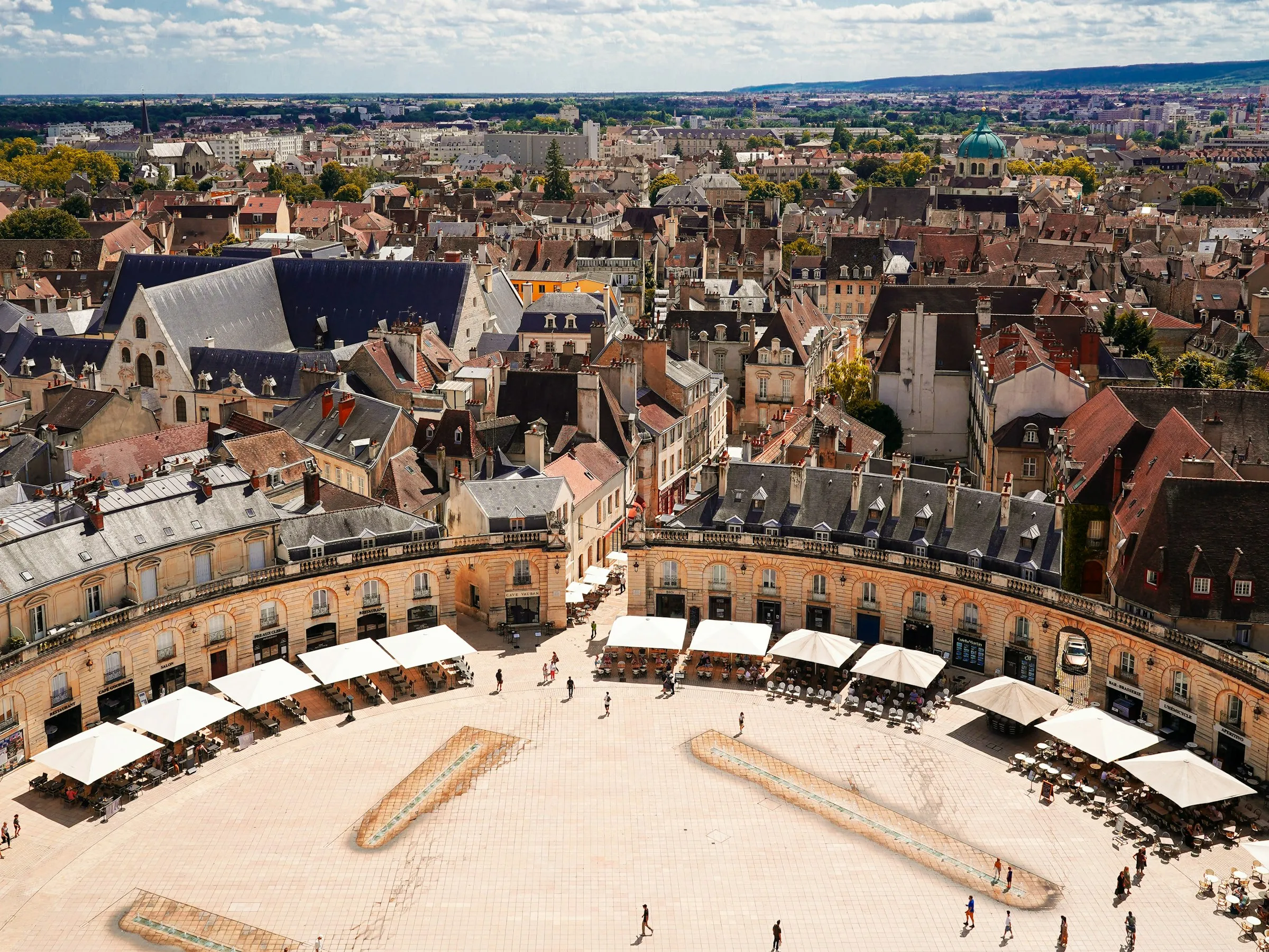Aerial view over Dijon's semicircular Place de la Liberation, curved arcaded facades and rows of cafe umbrellas ringing the pale stone plaza above red-tiled rooftops