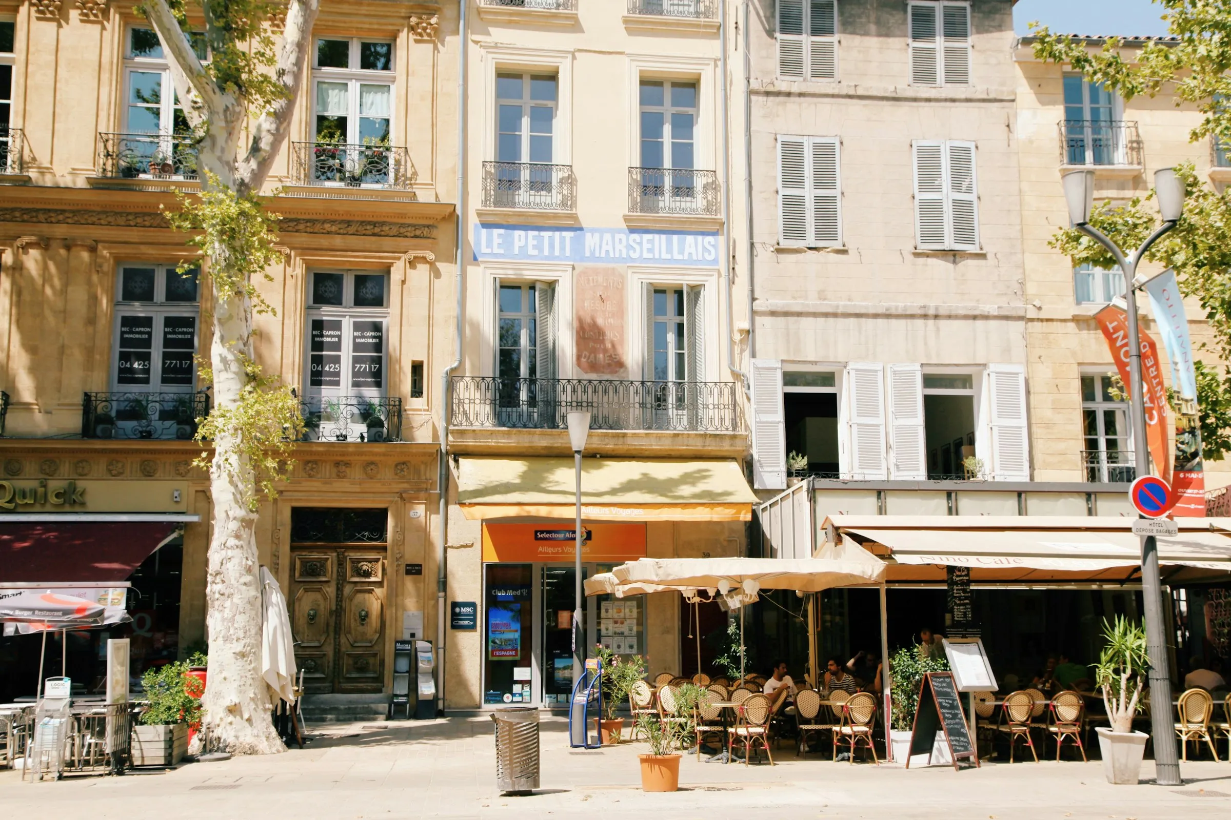 Cafe terraces with wicker bistro chairs under awnings along Aix-en-Provence's Cours Mirabeau, beneath tall plane trees and the historic painted 'Le Petit Marseillais' sign on a cream townhouse facade.