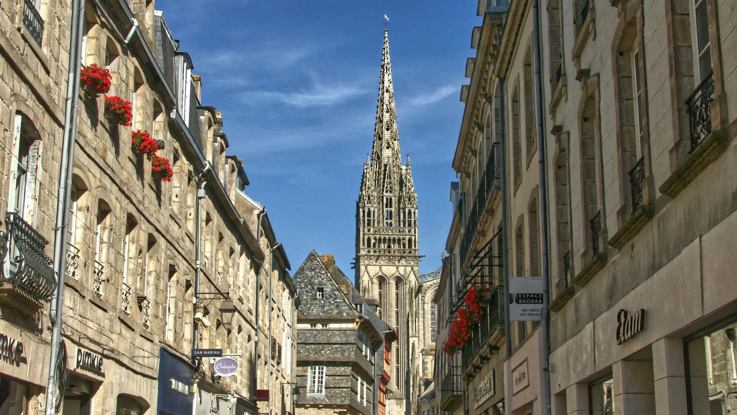 View up a narrow street of pale stone shopfronts hung with red geranium window boxes to the soaring openwork Gothic spire of Quimper's Cathedrale Saint-Corentin against a bright blue sky.
