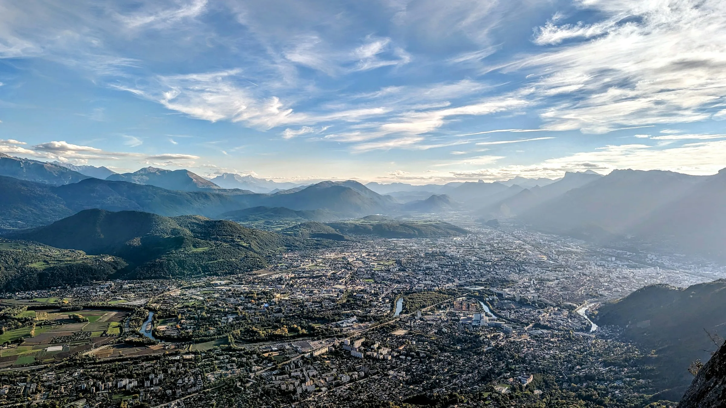 Golden-hour aerial panorama of Grenoble spread across the flat valley floor, completely ringed by layered blue Alpine ridgelines fading into haze, under a wide cloud-streaked sky.