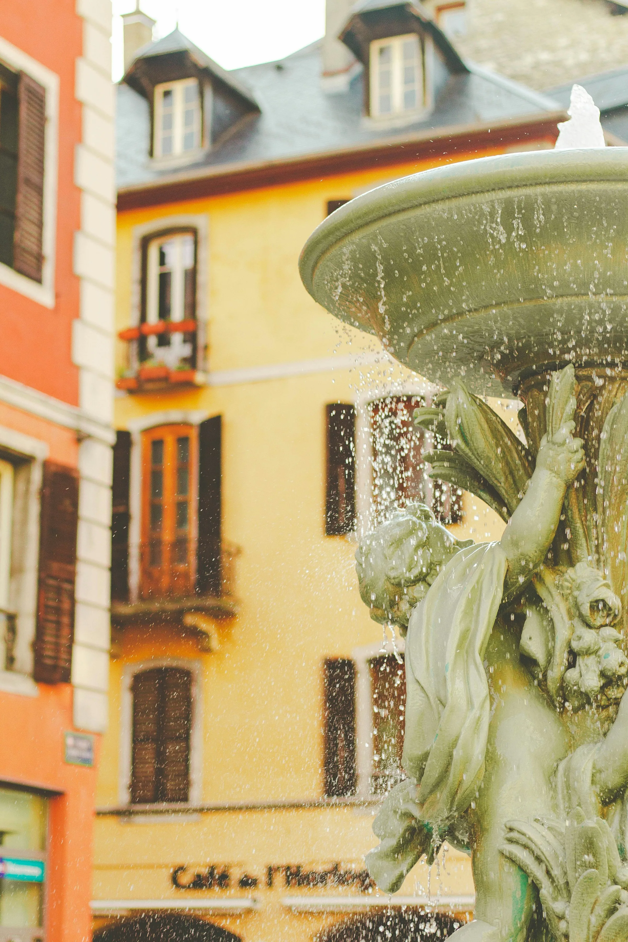 Close-up of Chambery's ornate Fontaine des Elephants, water spilling over its weathered bronze figures against the sunlit yellow and terracotta facades of the old town, with the Café de l'Horloge sign behind.