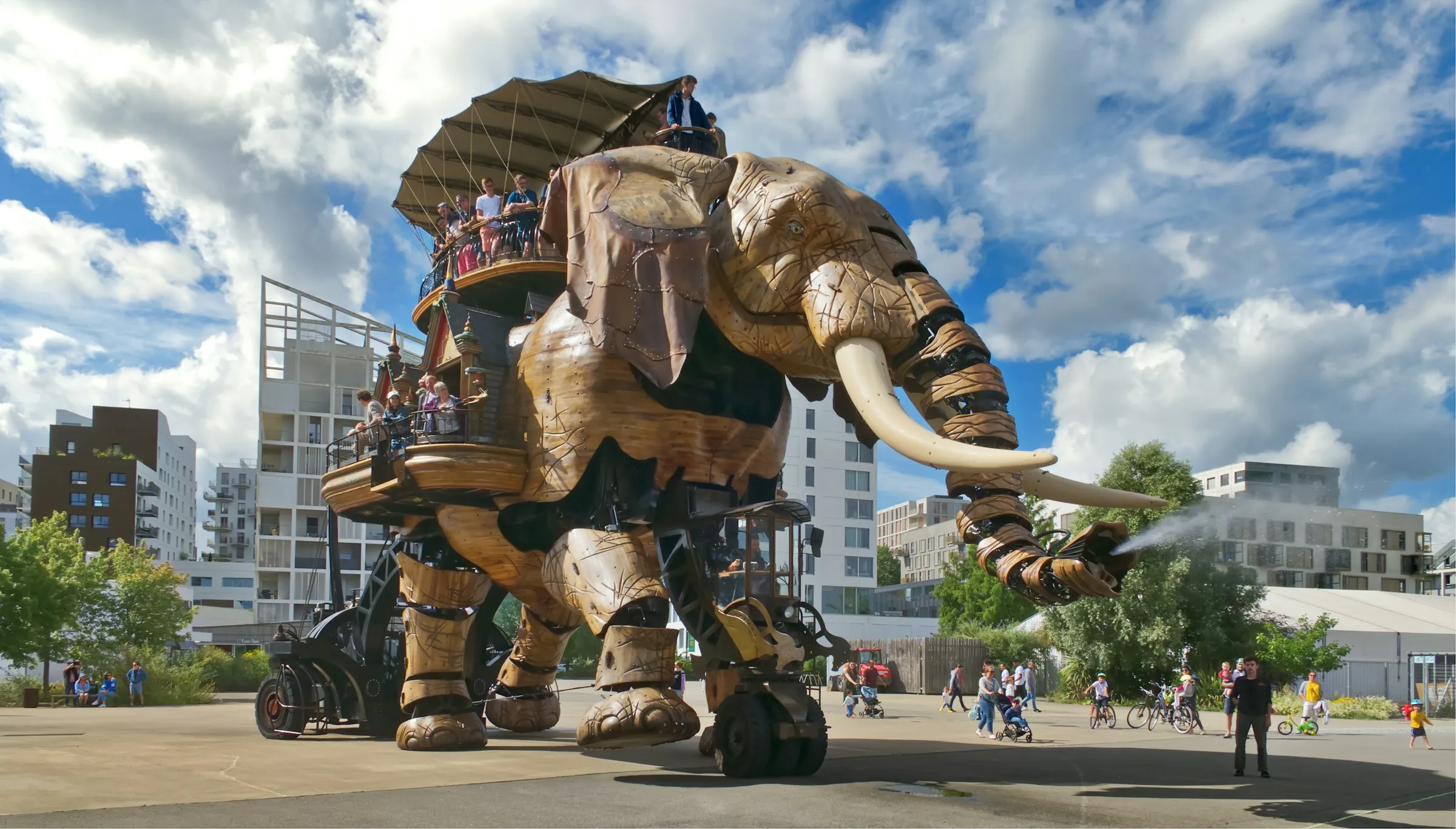 The Grand Elephant of the Machines de l'ile, a towering wood-and-steel mechanical elephant carrying riders on its back, strolls an open plaza in Nantes and sprays water from its trunk under a bright cloud-flecked sky.