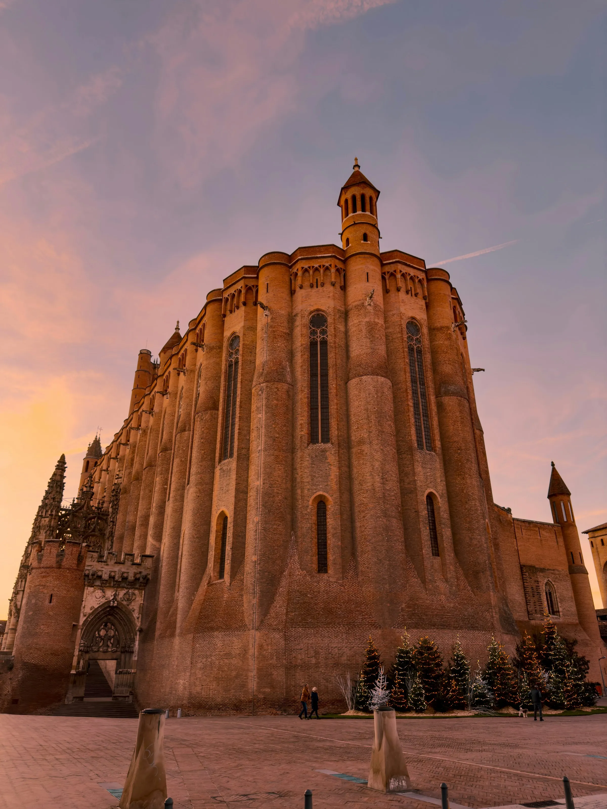 The fortress-like red-brick apse and octagonal turret of Cathedrale Sainte-Cecile in Albi glowing warm orange at golden-hour sunset under a pink sky, with lit Christmas trees along its base.