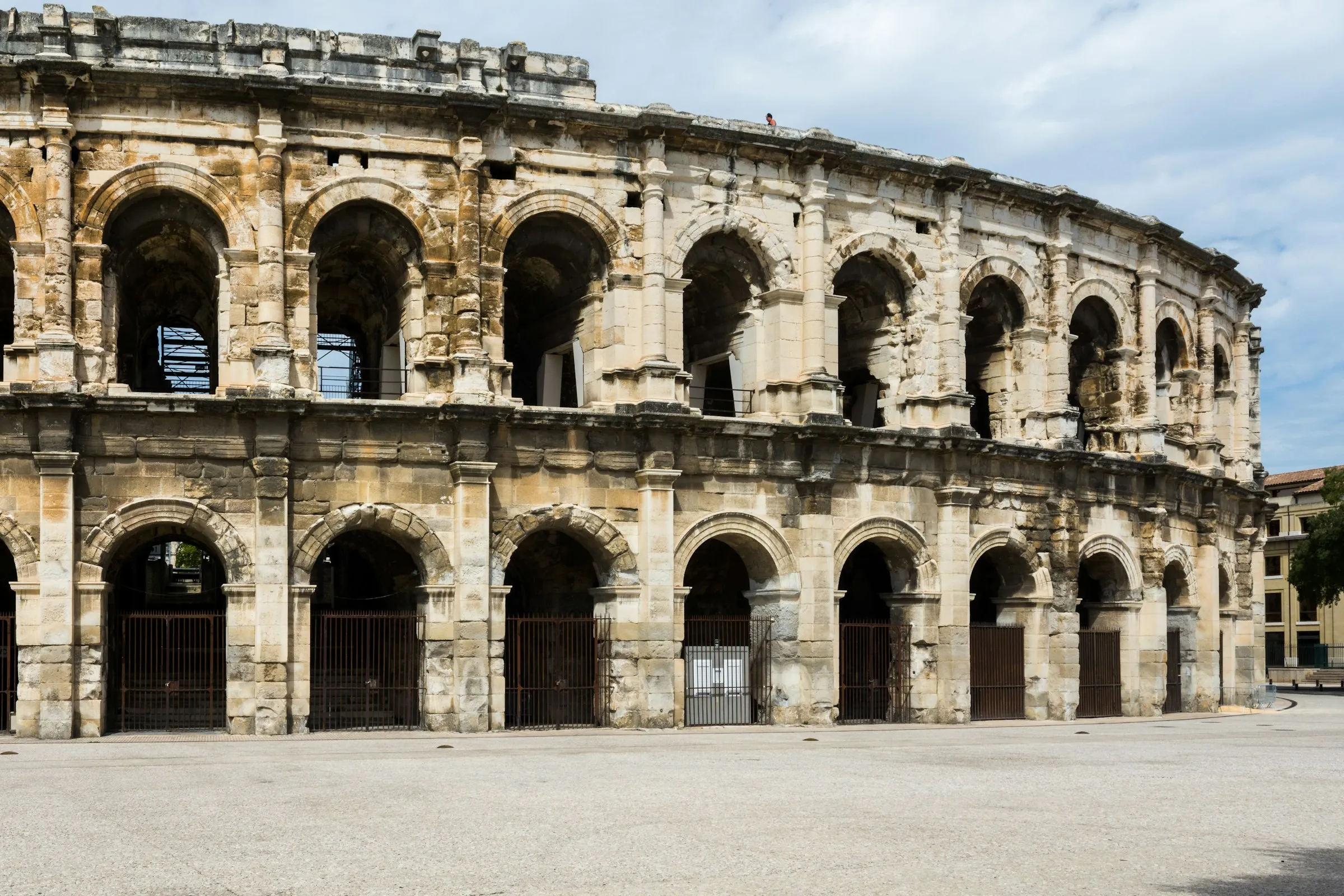 Close daylight view of the honey-colored limestone facade of the Arenes de Nimes, two superimposed rows of Roman arcades with engaged columns and the projecting attic corbels above, fronting an open sunlit square.