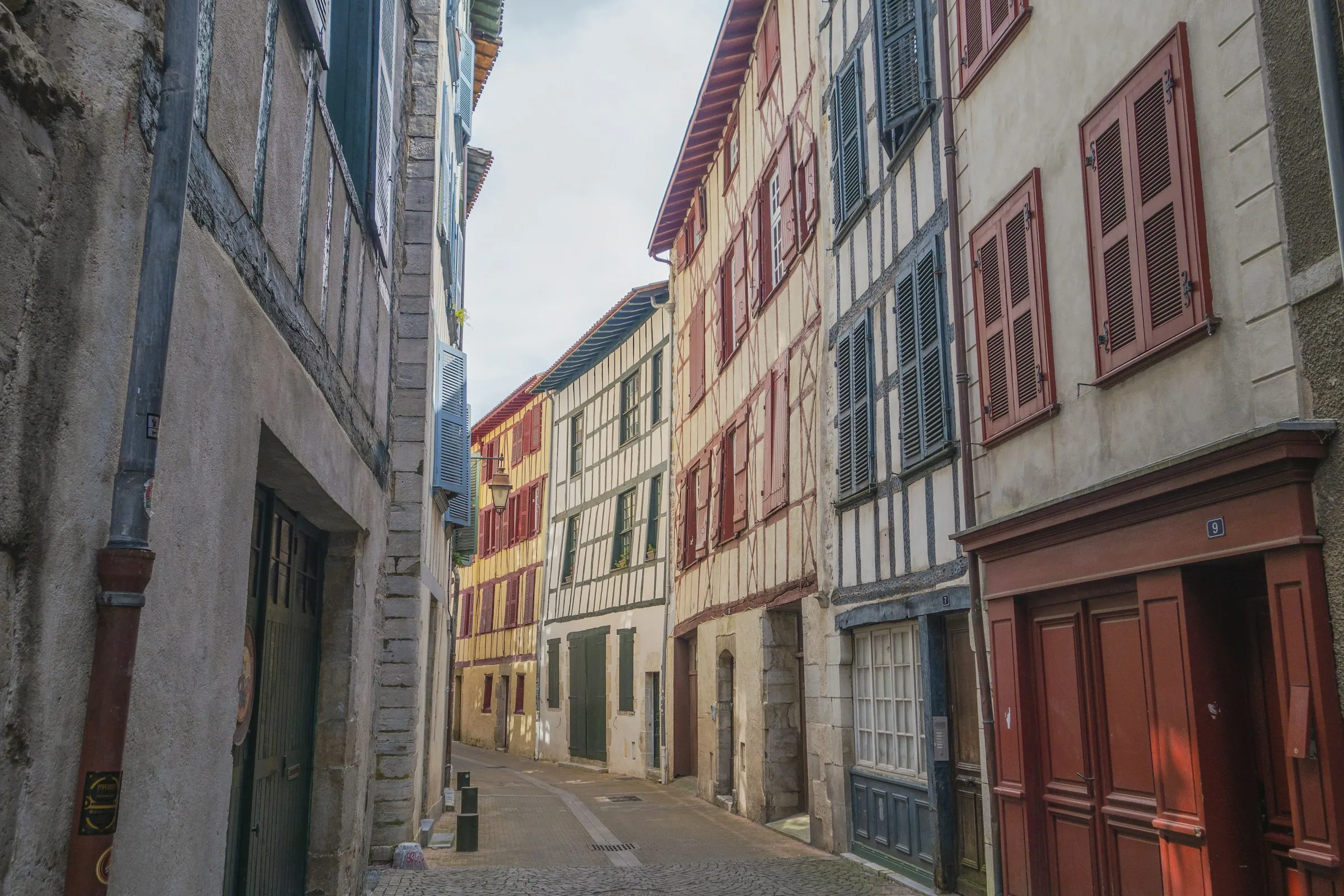 A quiet cobbled old-town street in Bayonne flanked by tall half-timbered Basque houses with red, blue and green shutters and painted timber framing.