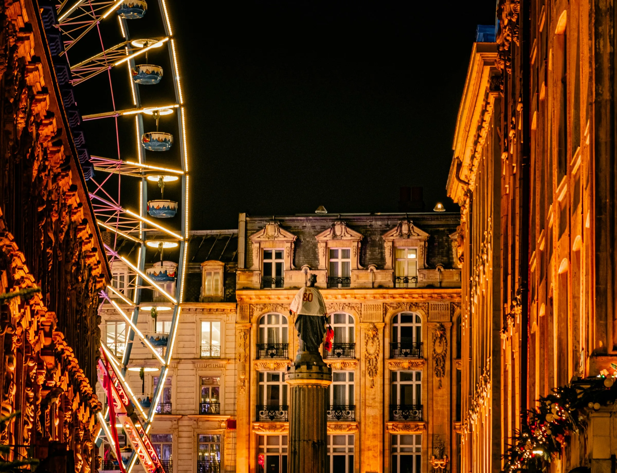 The Lille Christmas market at night on the Grand Place, an illuminated Ferris wheel glowing beside a warmly lit Haussmann-style building, with the Deesse statue on her column in the foreground.