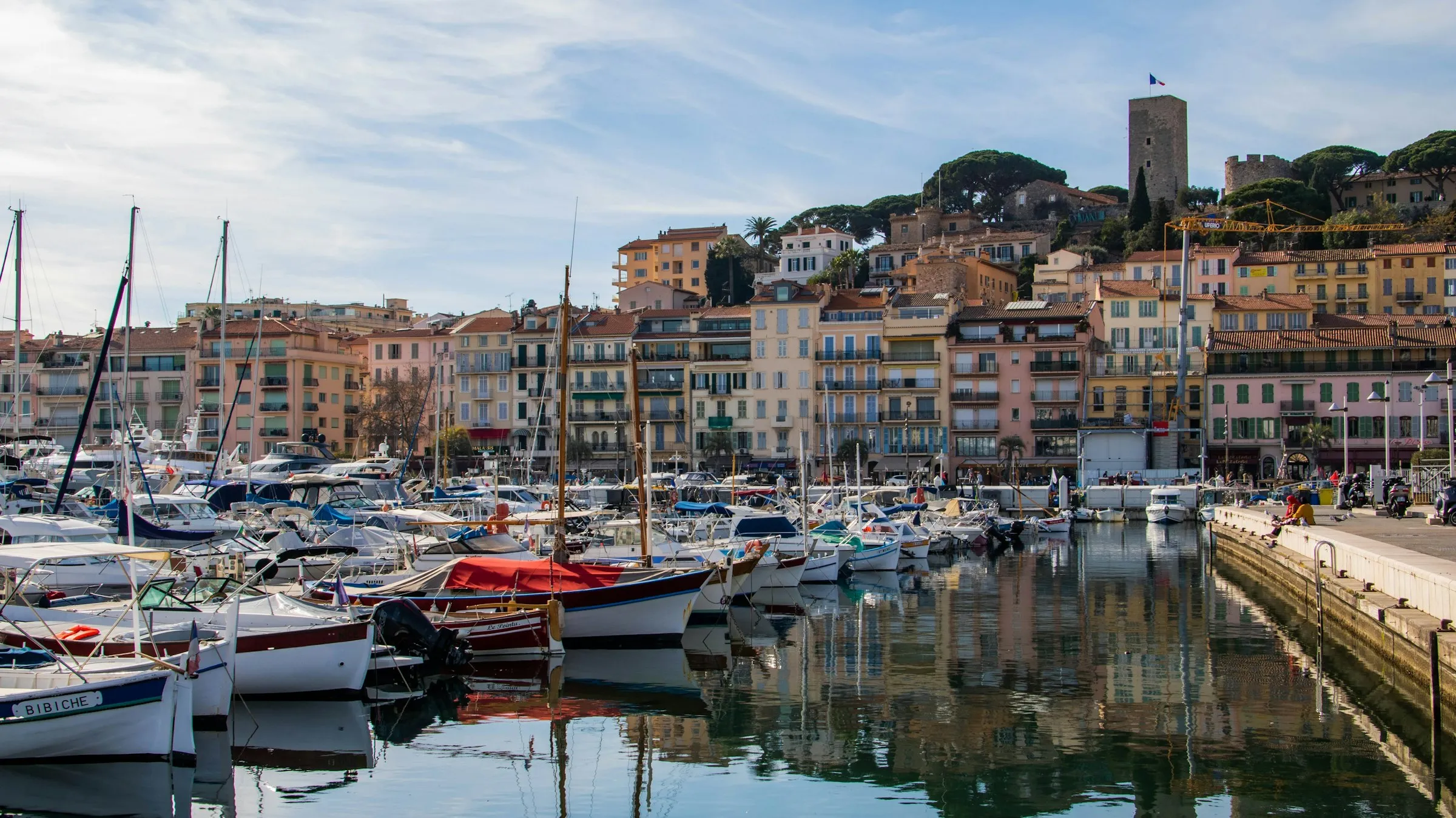 Cannes' Vieux-Port on a bright day, traditional wooden fishing boats moored along a pastel-coloured quay, with the medieval square tower and the Notre-Dame-d'Esperance clocktower rising on the Suquet hill behind.