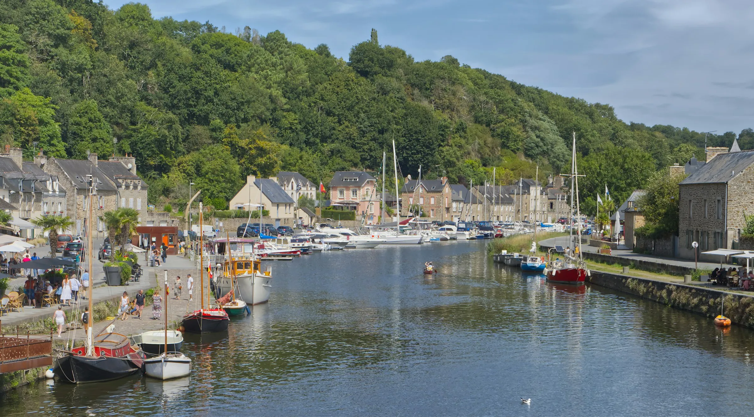 Sunny day at the Port de Dinan on the Rance, sailboats and cabin cruisers moored along the quay, waterfront cafe terraces with umbrellas and people strolling under a blue sky