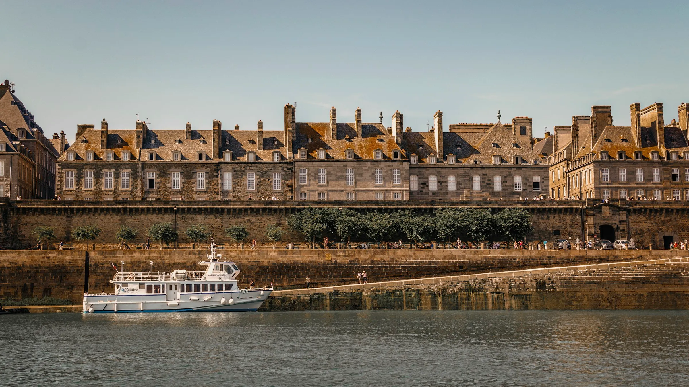 Tall granite townhouses with slate roofs and rows of chimneys rising behind Saint-Malo's stone ramparts, a white Corsaire ferry moored on the calm water below in golden late-afternoon light.