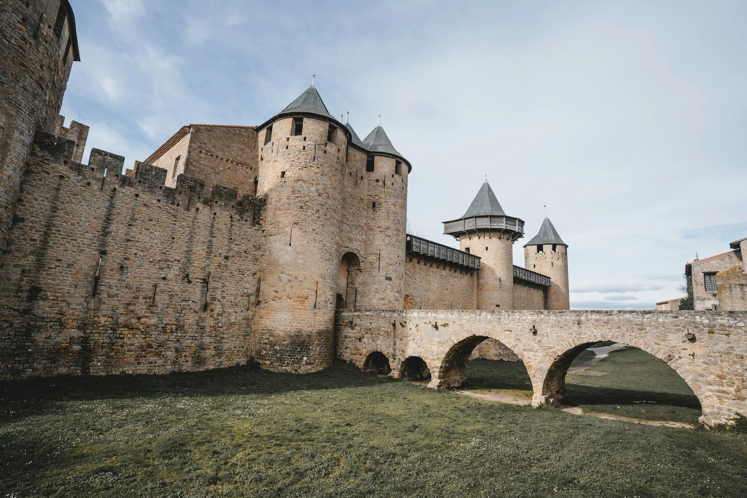 Low-angle view of the Chateau Comtal inside Carcassonne, round conical-roofed towers rising above an arched stone bridge over the grassy moat