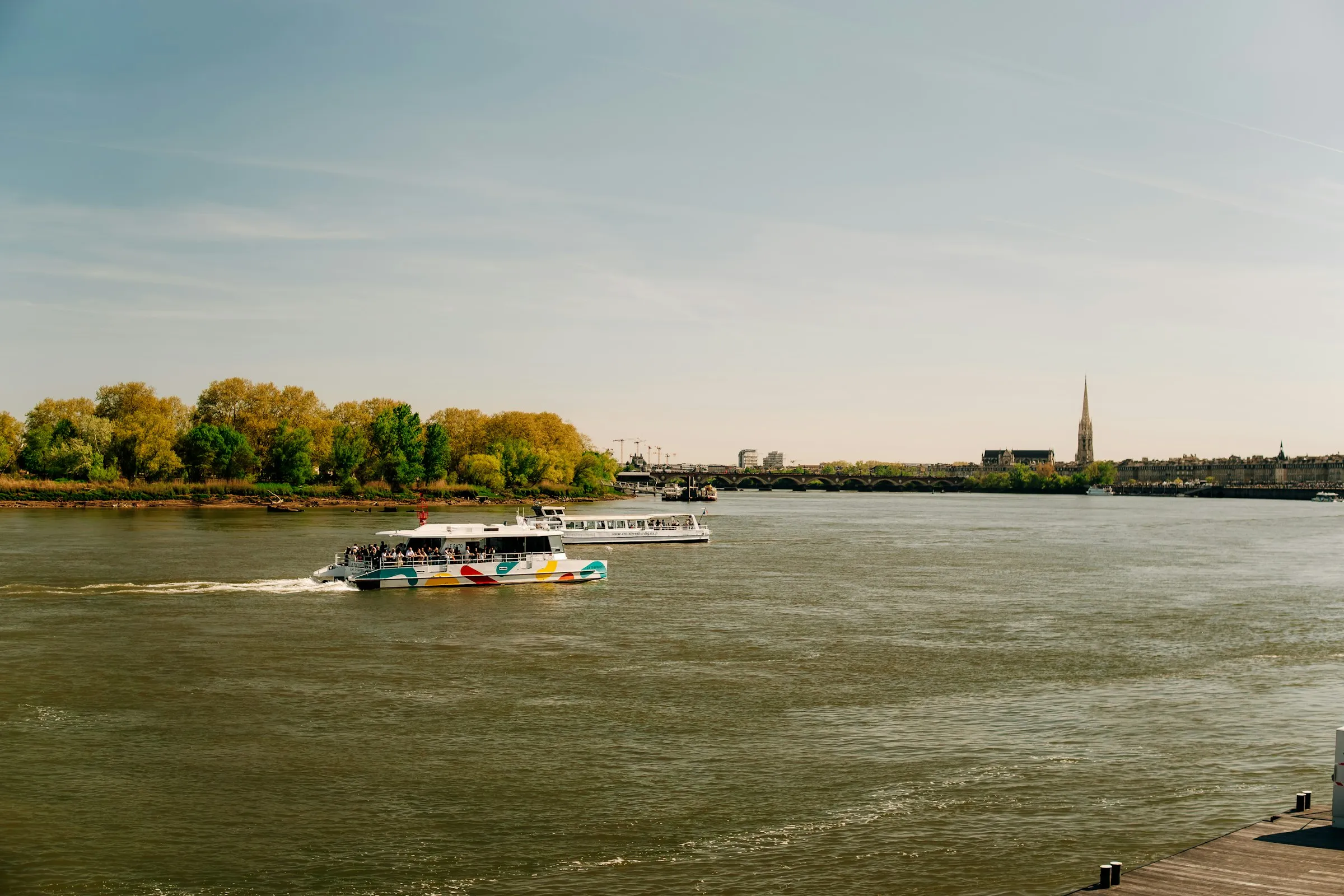 A colorful river shuttle and a tourist cruise boat moving across the wide sunlit Garonne at Bordeaux, the low stone arches of the Pont de Pierre and the tall spire of the Basilique Saint-Michel rising on the far bank.