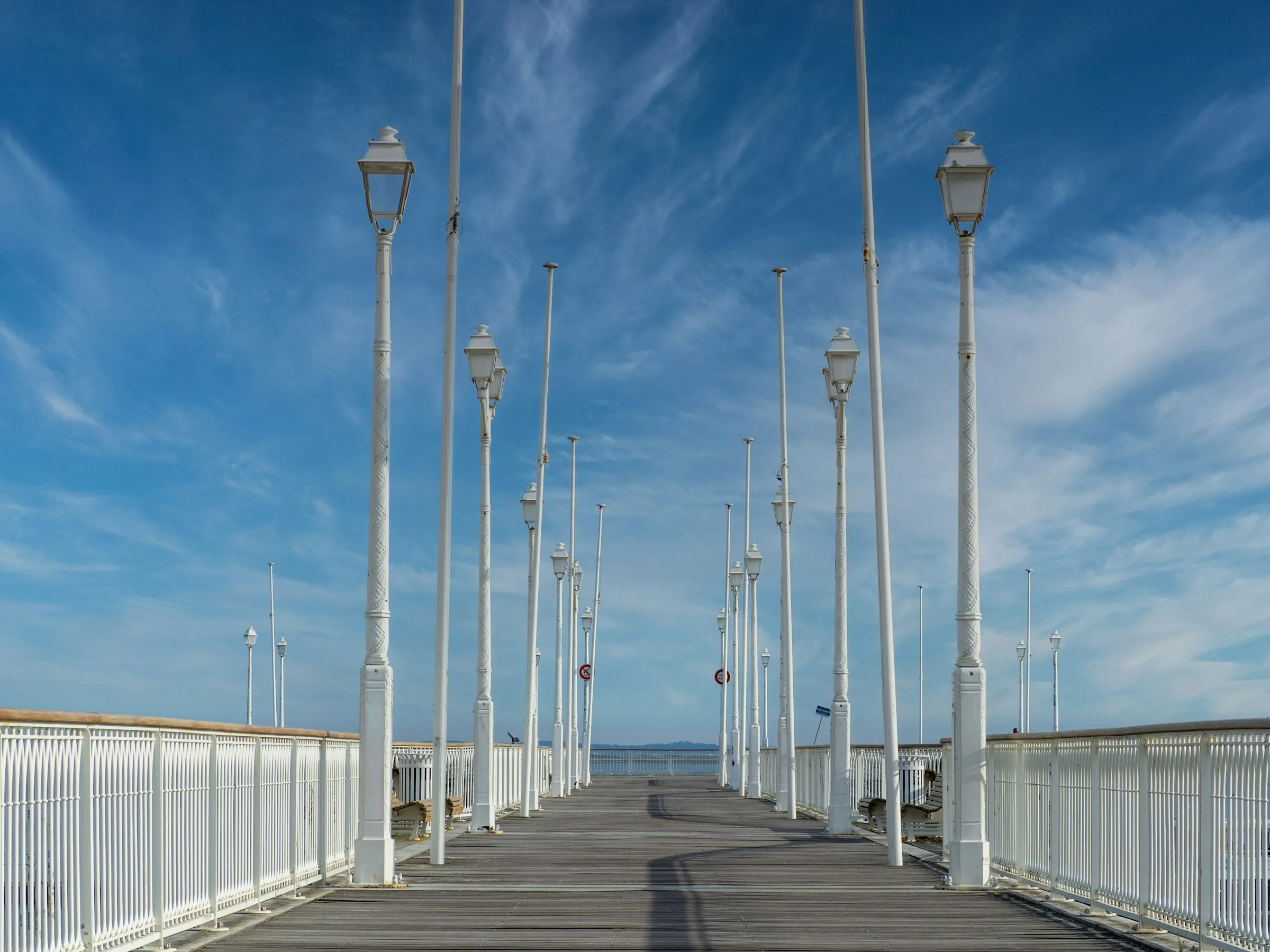 Arcachon's Belle Epoque pier stretching over the calm bay, lined with rows of ornate white cast-iron lampposts under a bright blue, wispy-clouded sky.