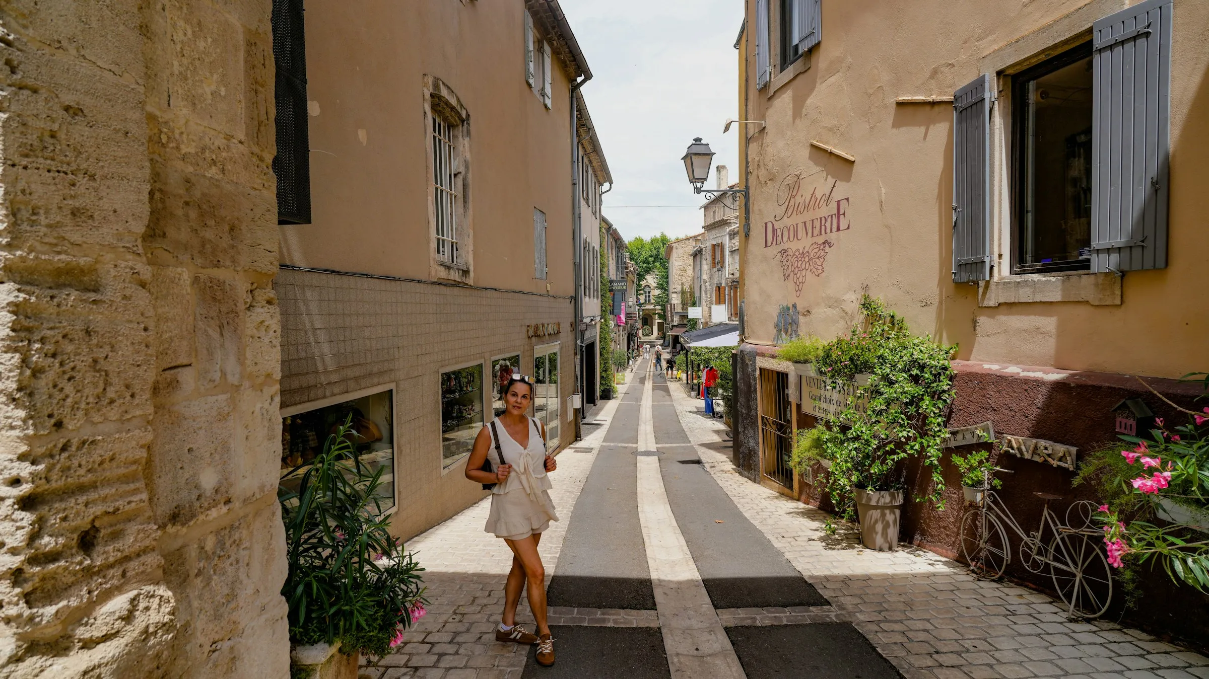 A sunlit cobbled lane in the old town of Saint-Remy-de-Provence, lined with ochre facades and grey-blue shutters, the hand-painted Bistrot Decouverte sign on the right and a woman strolling past.
