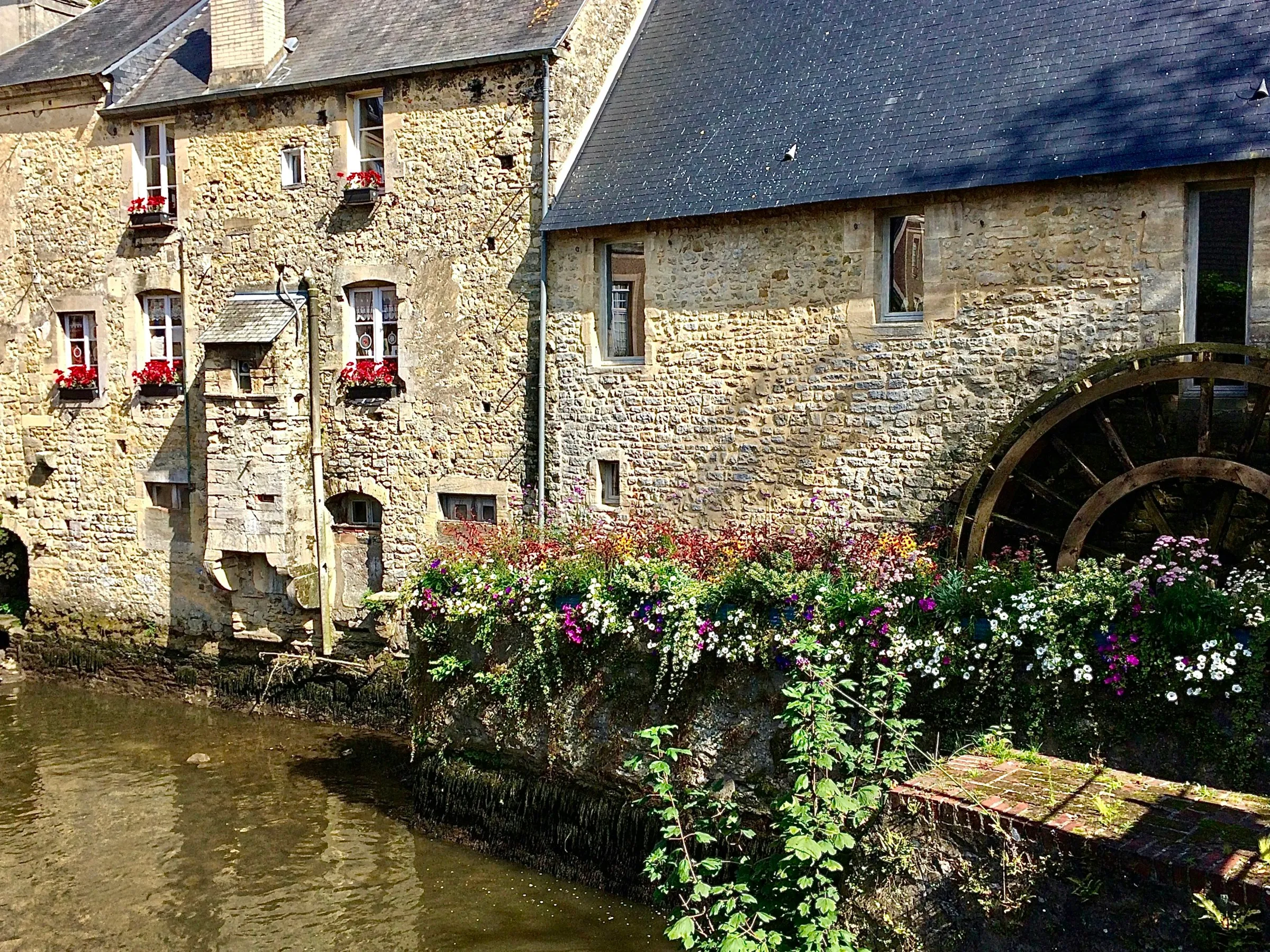 A honey-coloured stone watermill on the Aure in Bayeux, its old wooden waterwheel beside a bank spilling over with summer flowers, warm sunshine on the weathered stonework.