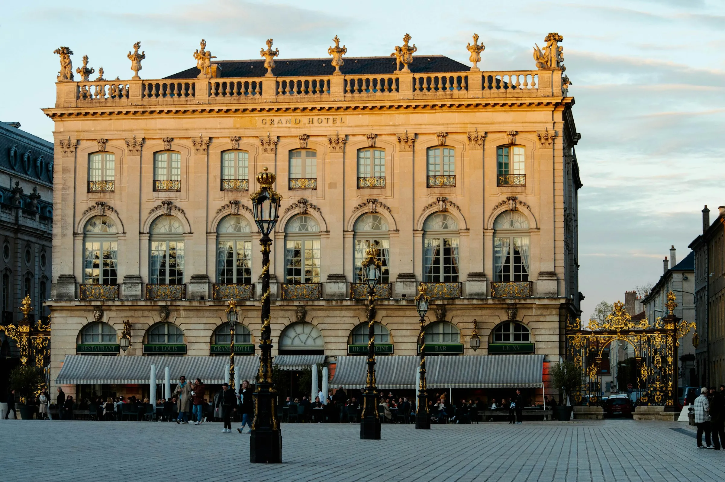 The honey-stone Grand Hotel facade on Place Stanislas glowing at golden hour, ornate gilded lamp posts and Jean Lamour ironwork gates in the foreground and diners seated at café terraces below the arched arcade