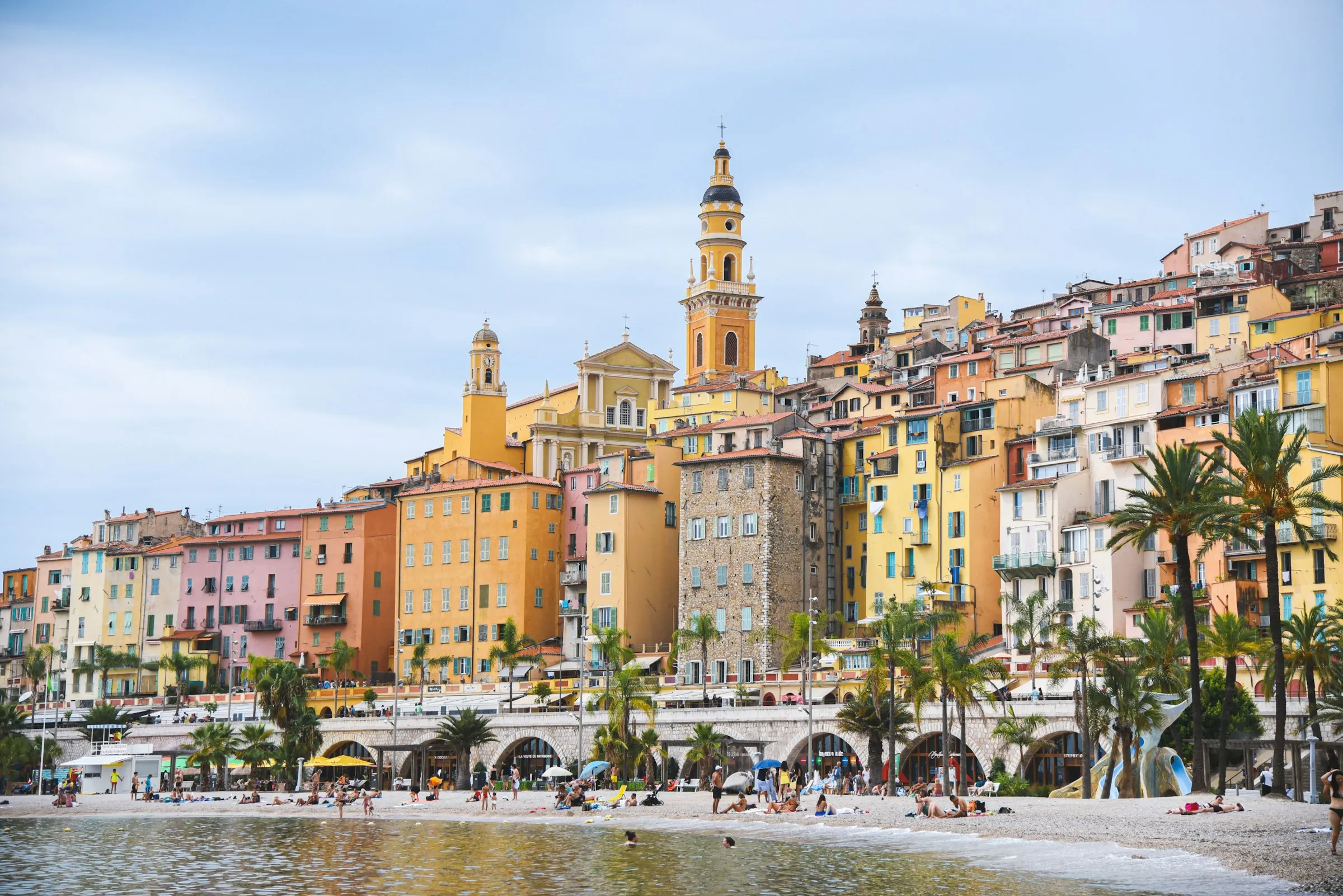 Menton's pebble beach on a bright sunny day, sunbathers and swimmers under palm trees below the tiered pastel old town crowned by the yellow Basilique Saint-Michel bell tower.