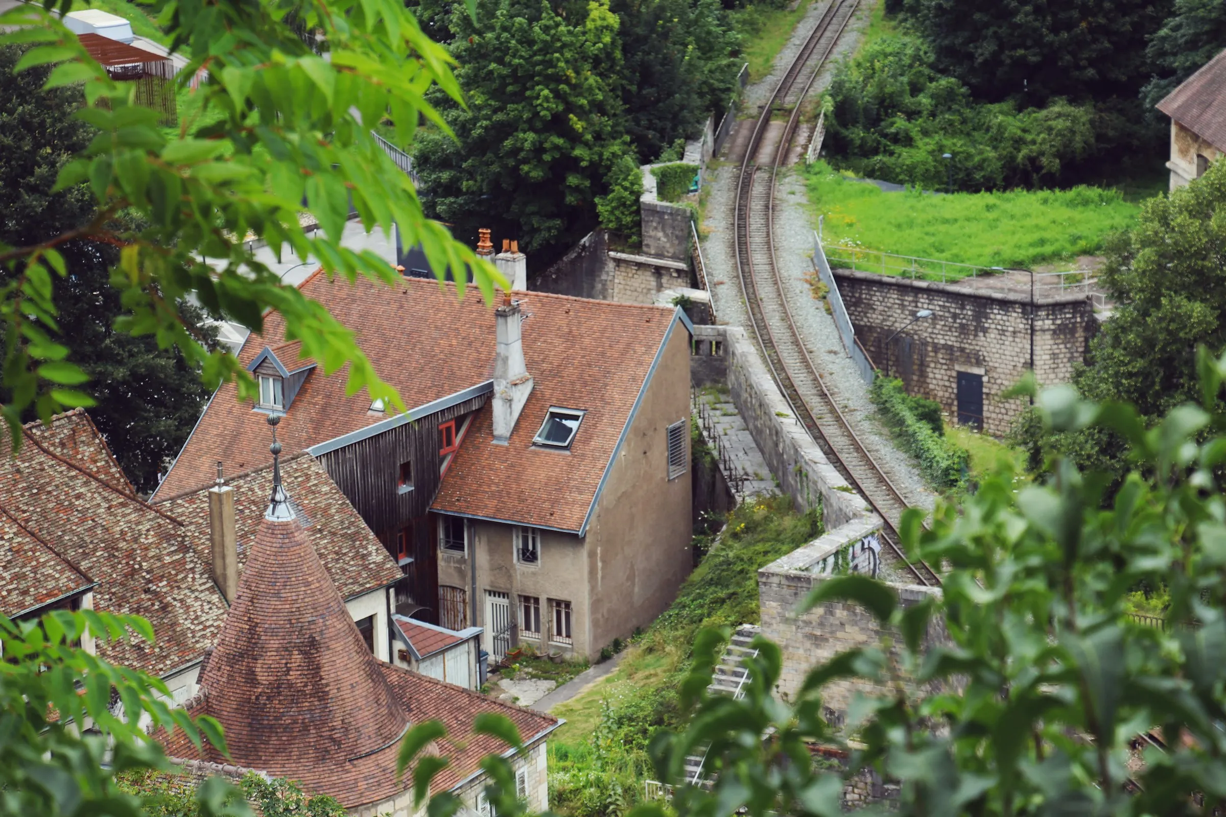 Elevated view over Besançon's honey-and-red tiled rooftops and a round pepperpot tower, with a single-track railway curving away along a lush green wooded hillside.