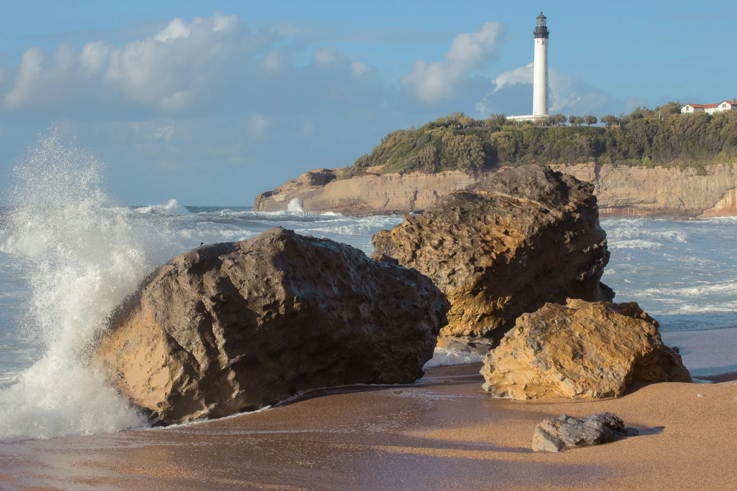 Golden evening light on a Biarritz beach with large sea-worn boulders on the sand, surf breaking behind them, and the white Phare de Biarritz lighthouse on the clifftop beyond