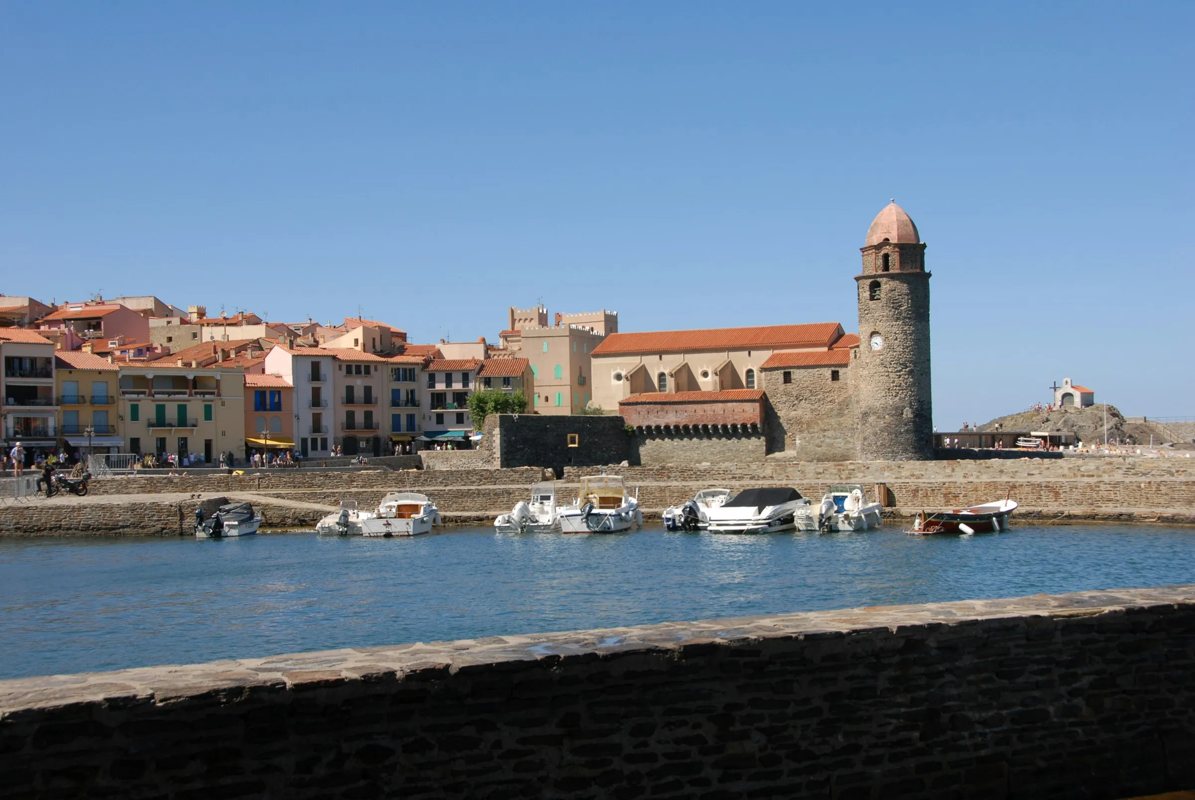 The harbour at Collioure on a clear blue-sky day, small boats moored in front of the pastel Catalan houses and the landmark pink-domed bell tower
