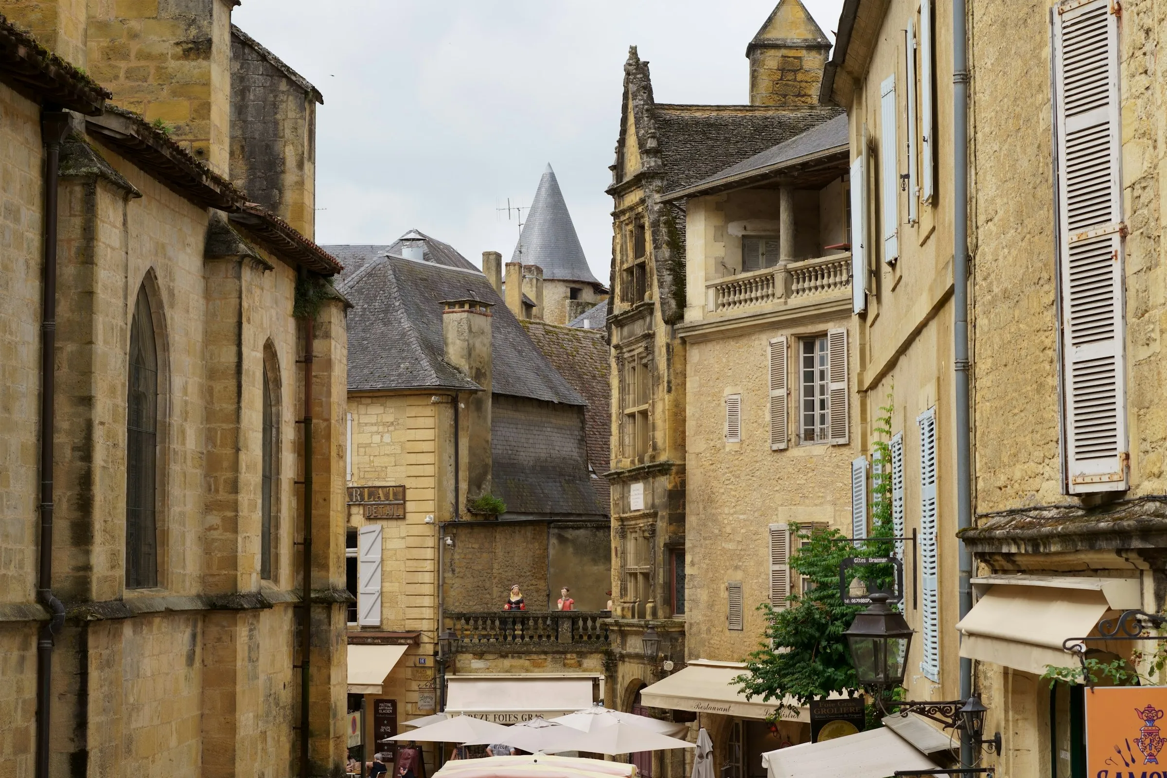 An elevated view over Sarlat-la-Caneda's honey-stone rooftops and a pointed lauze-roofed turret, a Gothic church on the left and market umbrellas with foie gras stalls in the lane below