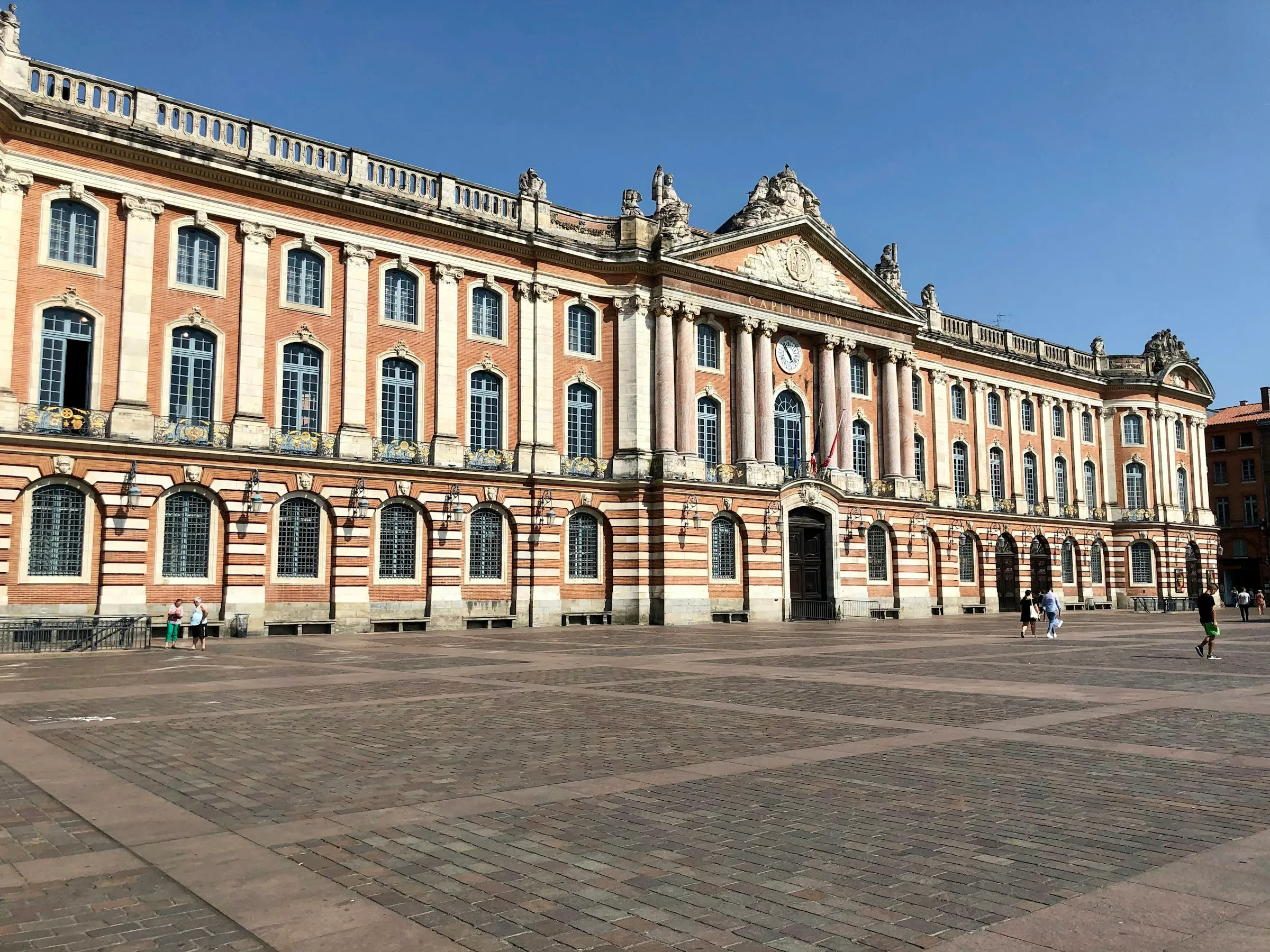 The long pink-brick and pale-stone facade of the Capitole de Toulouse under a clear blue sky, its columned central pavilion and CAPITOLIUM inscription facing the wide paved square.