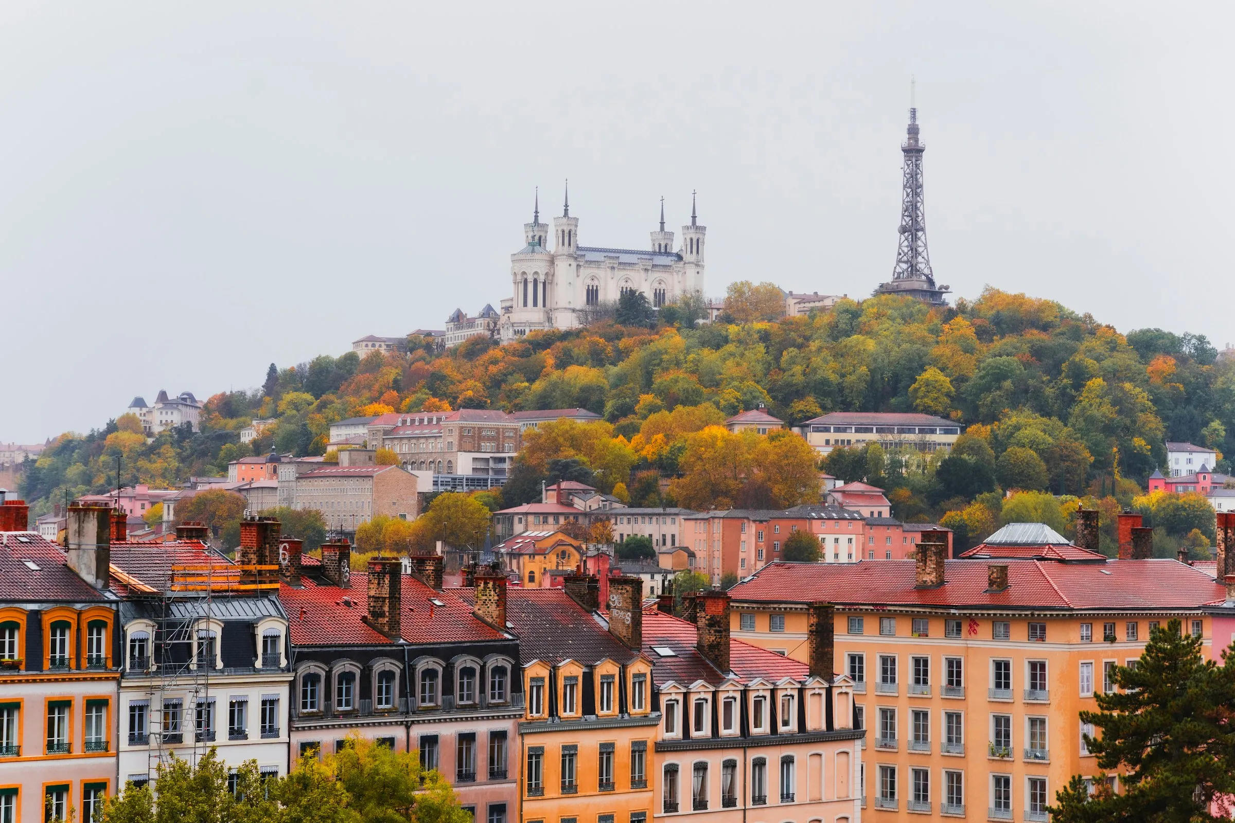 Lyon's hillside in autumn: the Fourviere basilica and metal tower rising above red-tiled rooftops and orange-and-gold trees, with warm-toned apartment facades in the foreground