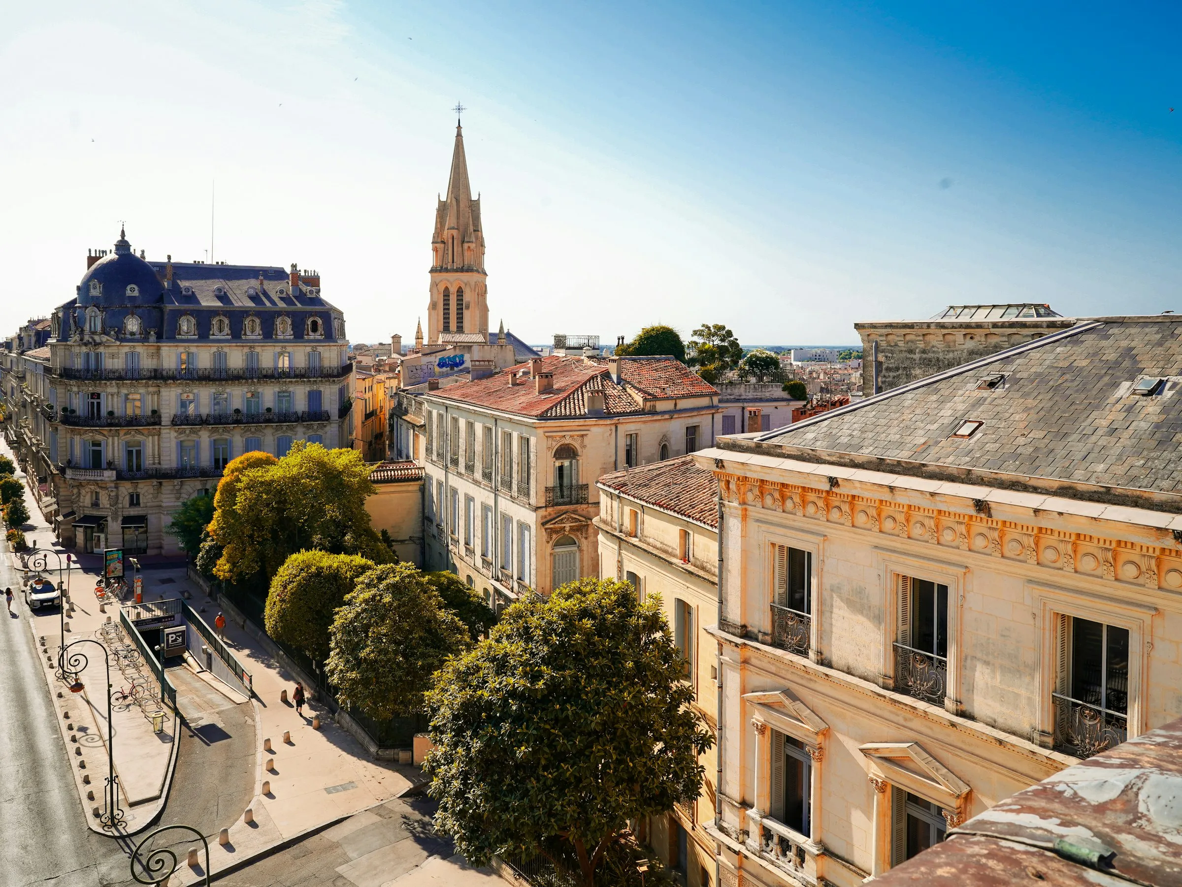 Sun-warmed rooftops of Montpellier's Ecusson old town, with a Haussmann-era slate dome and the Gothic spire of the Sainte-Anne church rising against a bright blue sky.