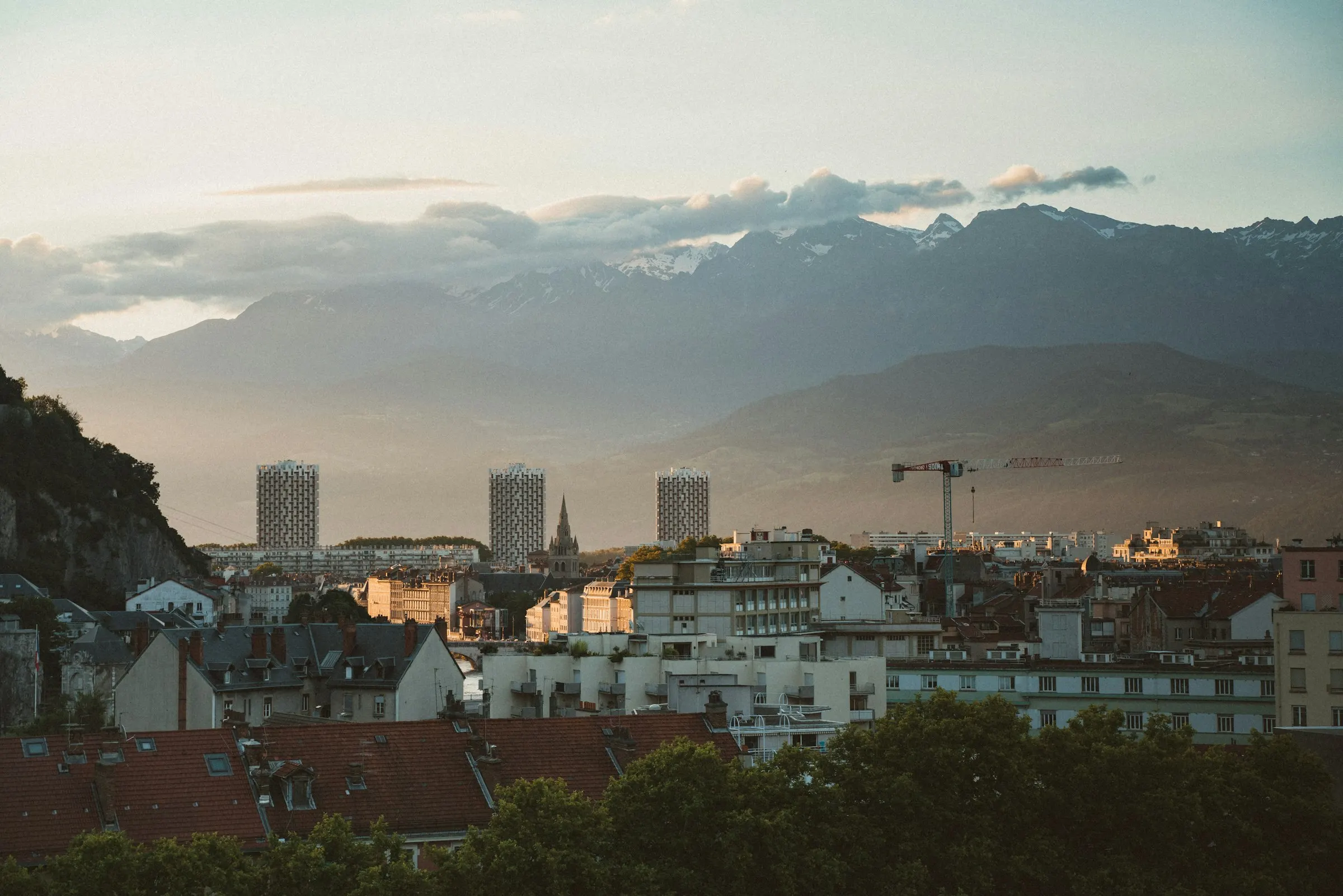 Grenoble at sunset seen across warm red-tiled rooftops, the three tall Île Verte towers and a slender church spire in the mid-ground, with the snow-capped Belledonne range glowing behind.