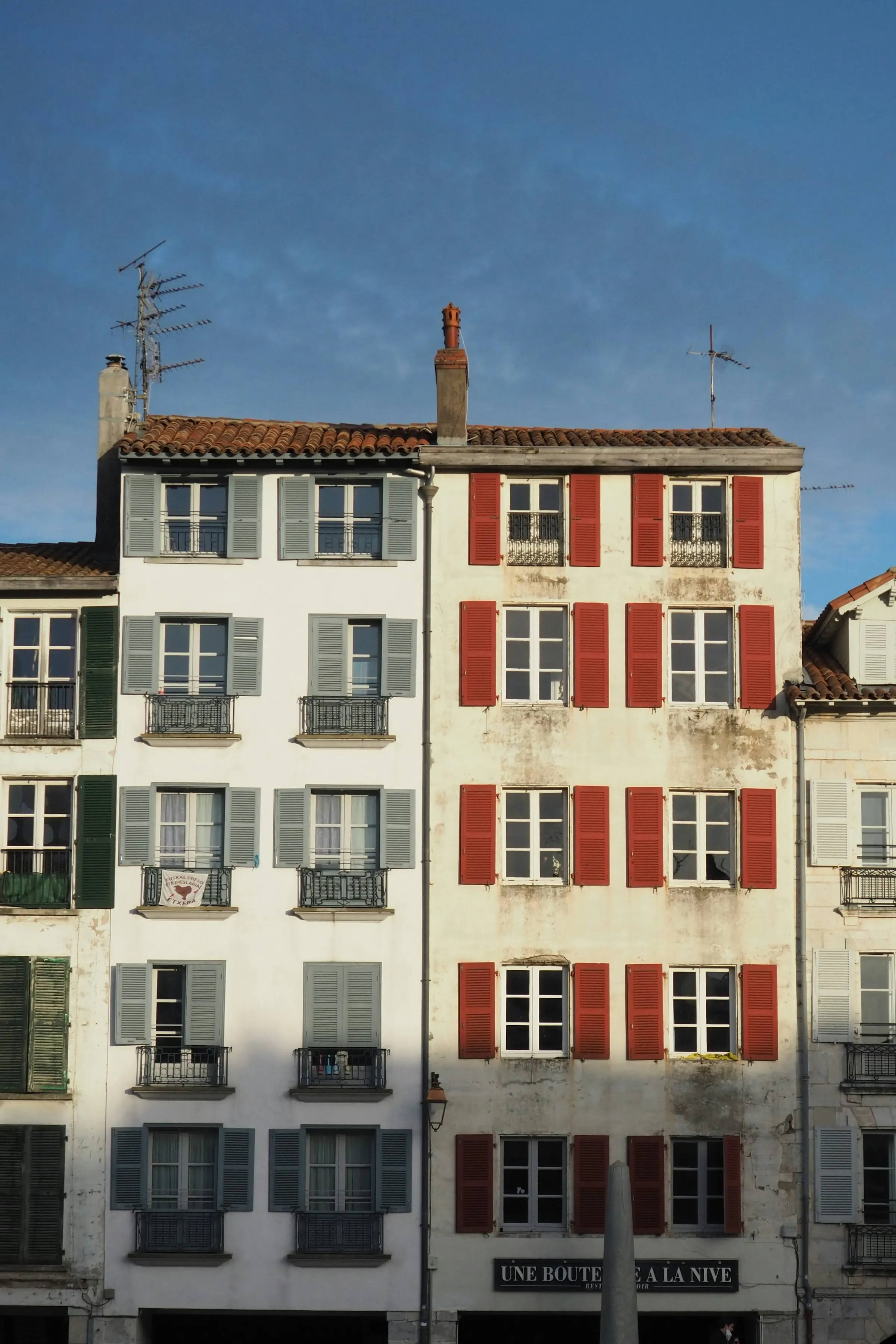 Warm golden light on tall Basque facades with red and grey-green shutters and wrought-iron balconies above the 'Une Bouteille a la Nive' restaurant in Bayonne's old town, against a clear blue sky.