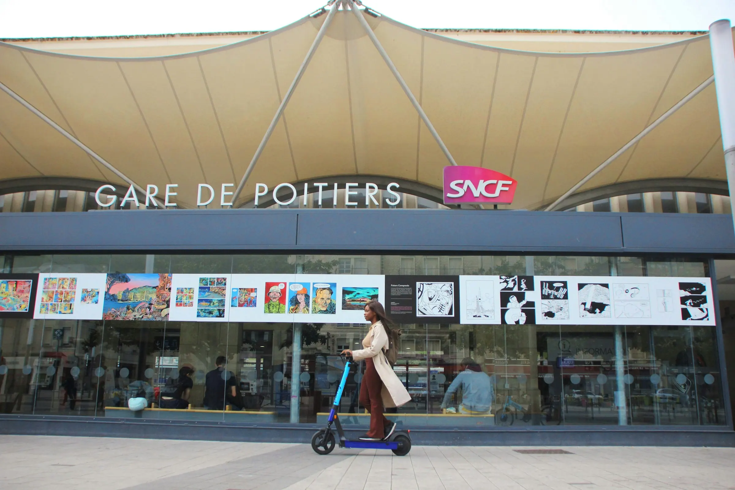 A woman rides an e-scooter past the sweeping tent-canopy entrance of the Gare de Poitiers SNCF station on a bright day, the city's direct TGV link toward Paris.