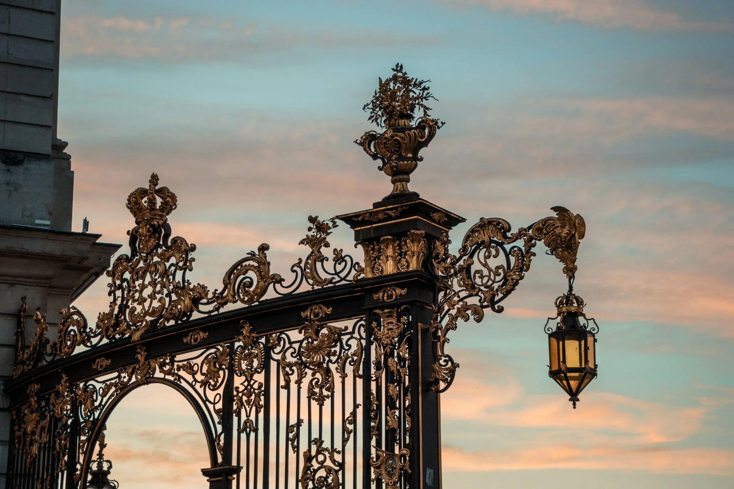 Close-up of Nancy's celebrated gilded wrought-iron gates by Jean Lamour, with rococo scrollwork, a golden crown and flowered urn and a hanging lantern silhouetted against a soft pink sunset sky