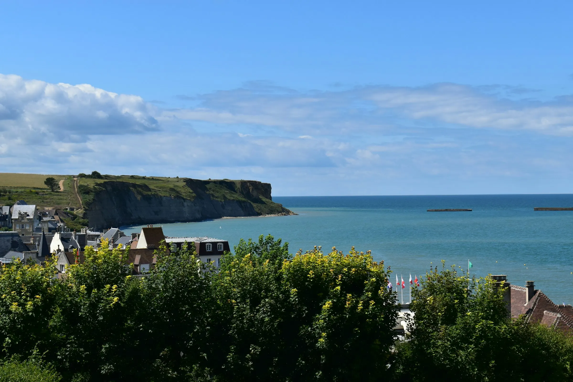 A sunny view over Arromanches-les-Bains on Normandy's D-Day coast near Caen — a turquoise bay framed by a green headland, red-tiled seaside rooftops and the concrete Mulberry-harbour caissons still dotting the water offshore