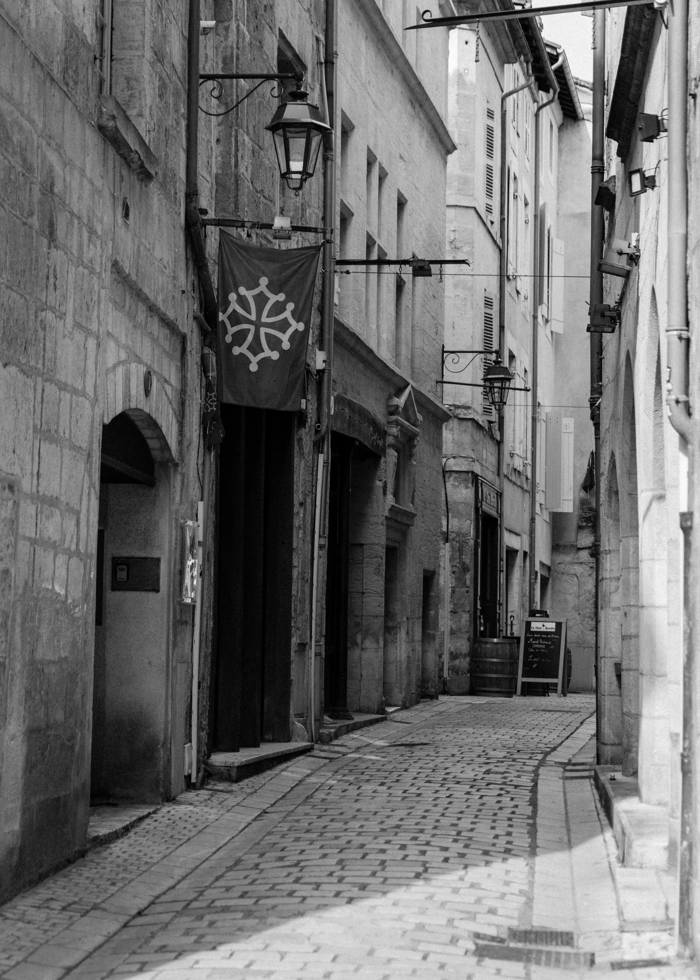 A narrow sunlit cobbled street in the old town, an Occitan-cross banner hanging from a stone facade and a cafe's chalkboard menu set out on the pavement