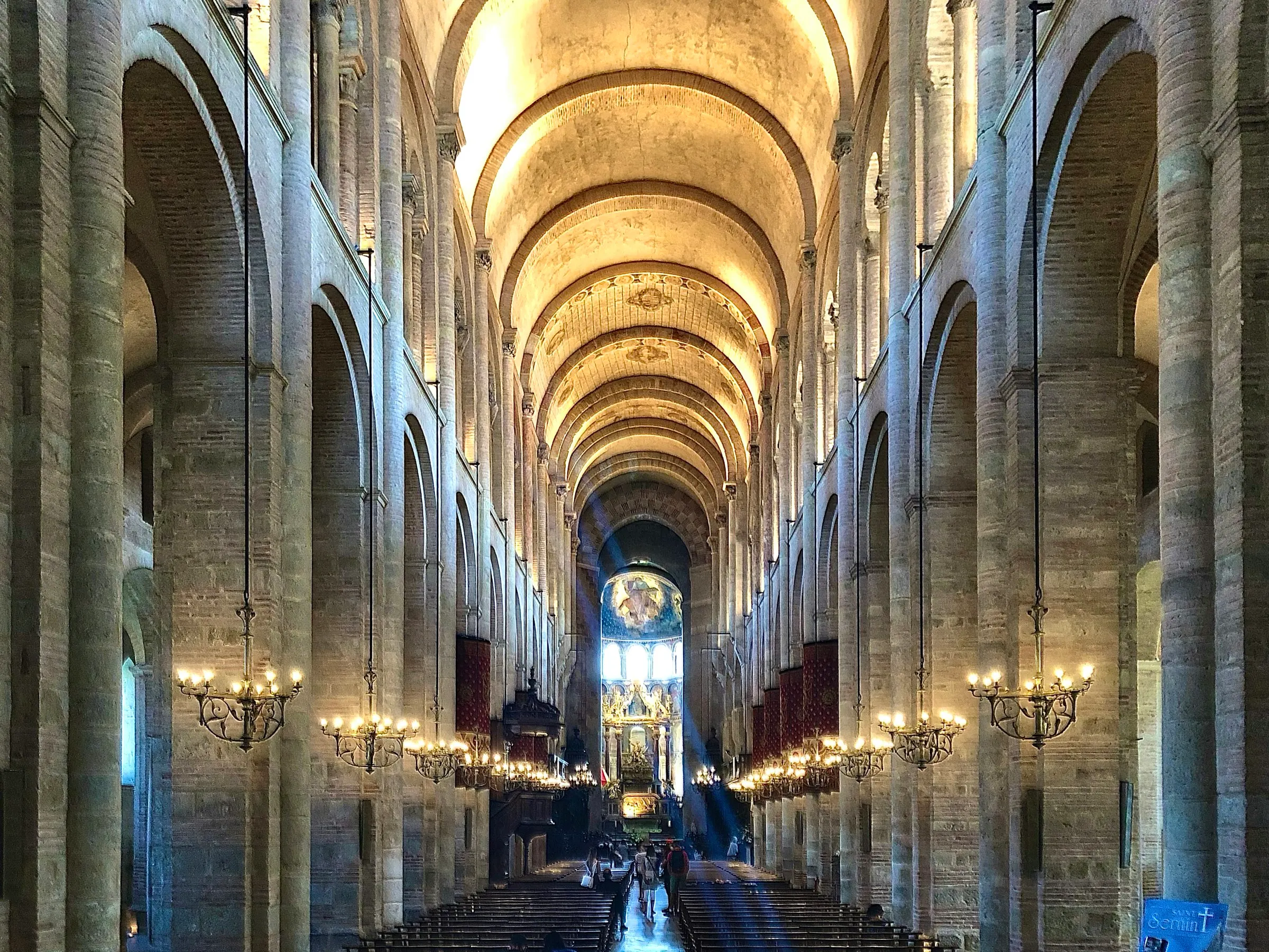 The vast Romanesque nave of the Basilica of Saint-Sernin in Toulouse, warm brick barrel vaulting and rows of hanging chandeliers, a shaft of daylight cutting across the aisle toward the golden apse.