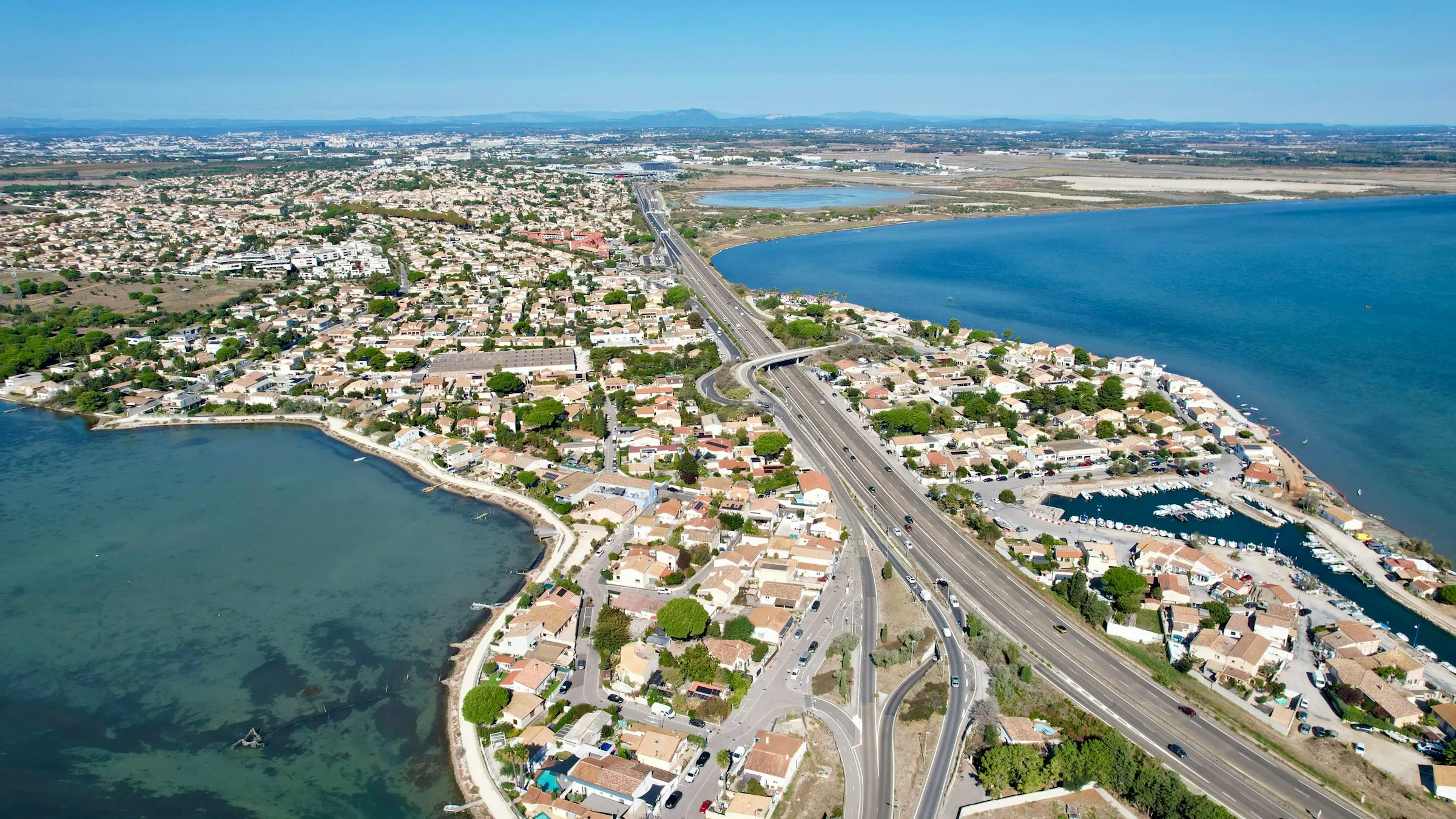 Aerial view of the residential Mediterranean coast just outside Montpellier, where seaside neighborhoods and a small marina thread between a blue lagoon and the open sea.