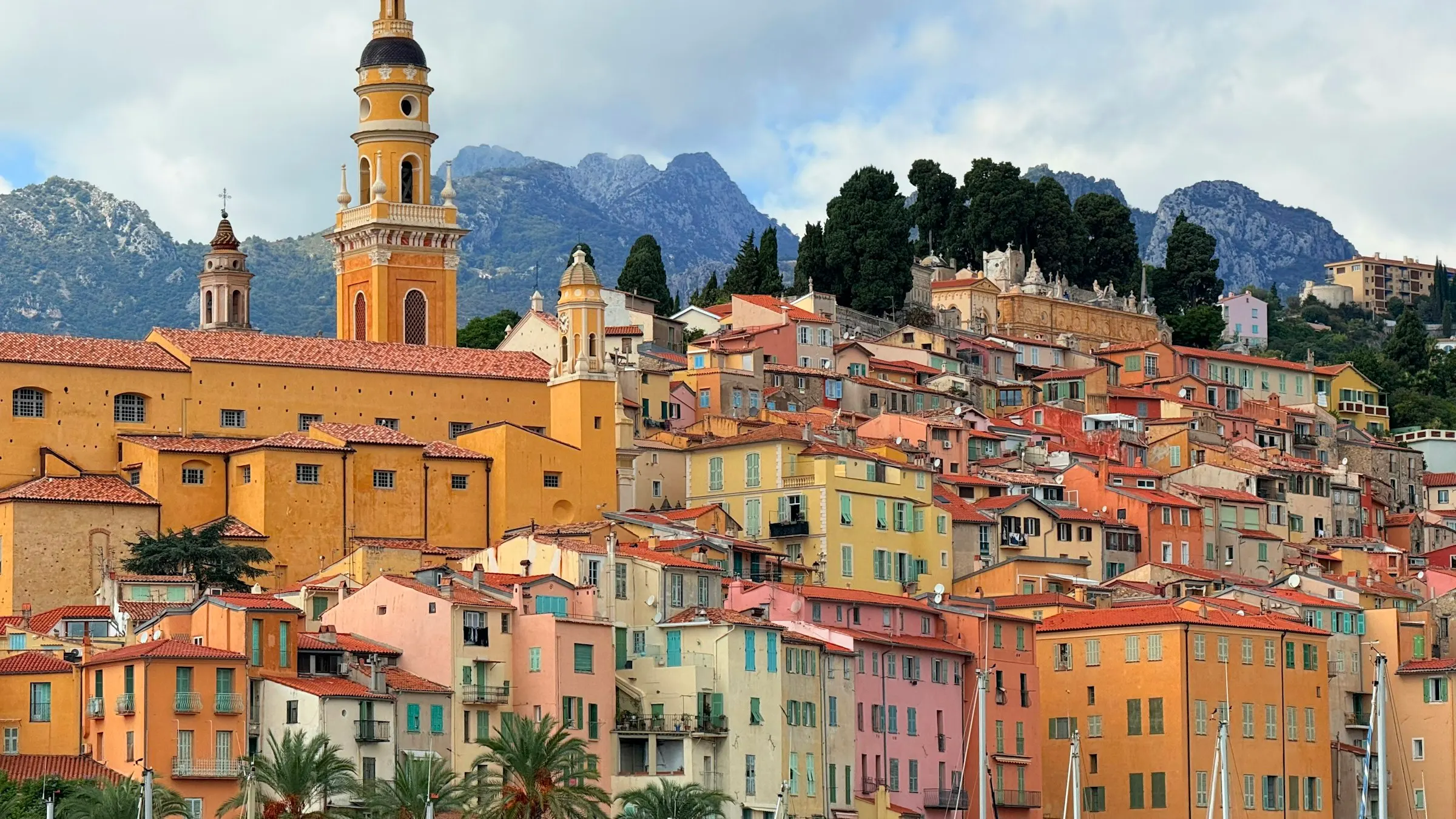 Close-up of Menton's old town: brightly painted ochre, pink and yellow houses stacked up the hillside beneath the ornate yellow Saint-Michel bell tower, with mountains rising behind.