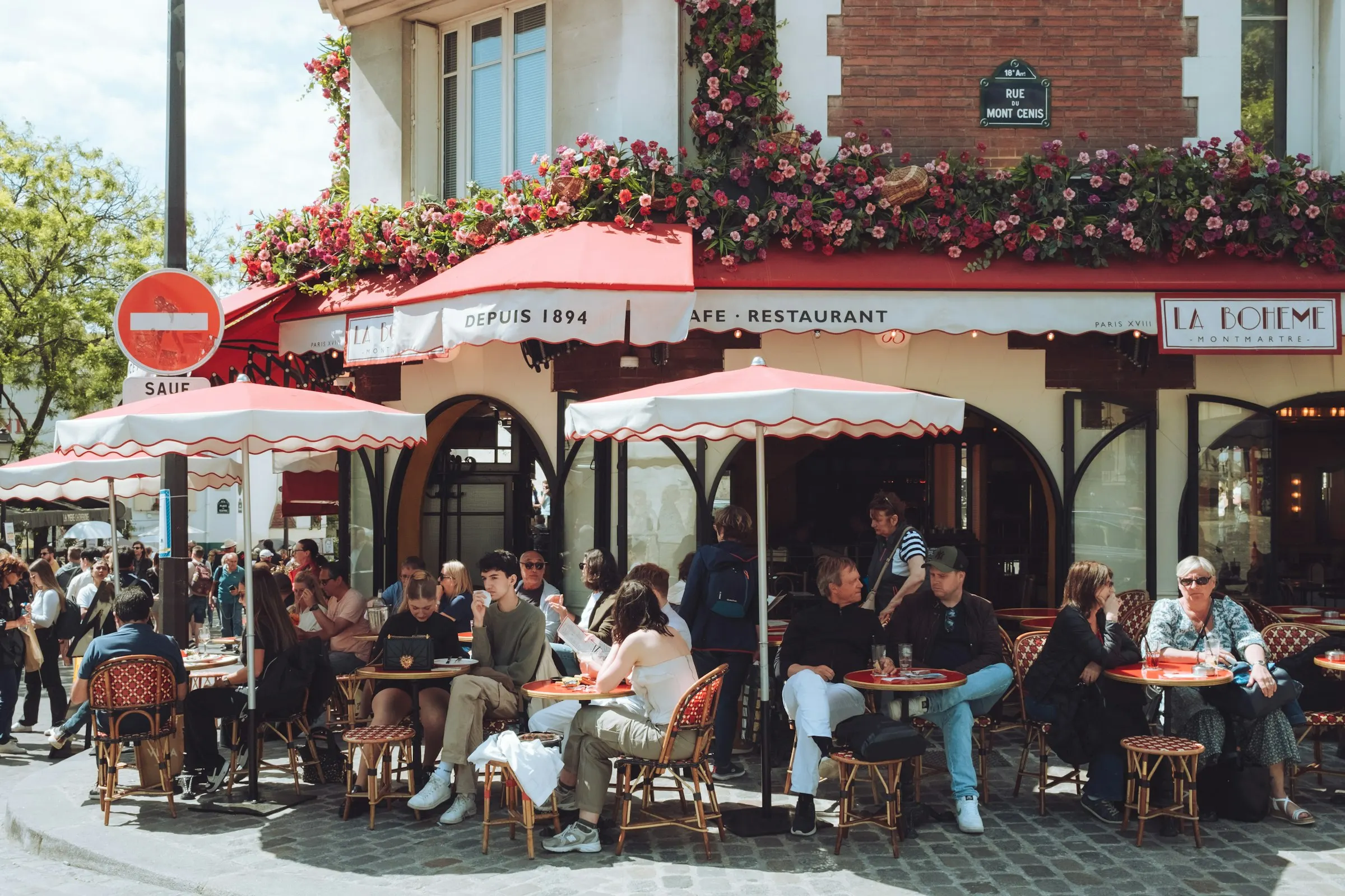 Parisians relaxing under red awnings at the flower-draped La Bohème café terrace on the Rue du Mont Cenis in Montmartre