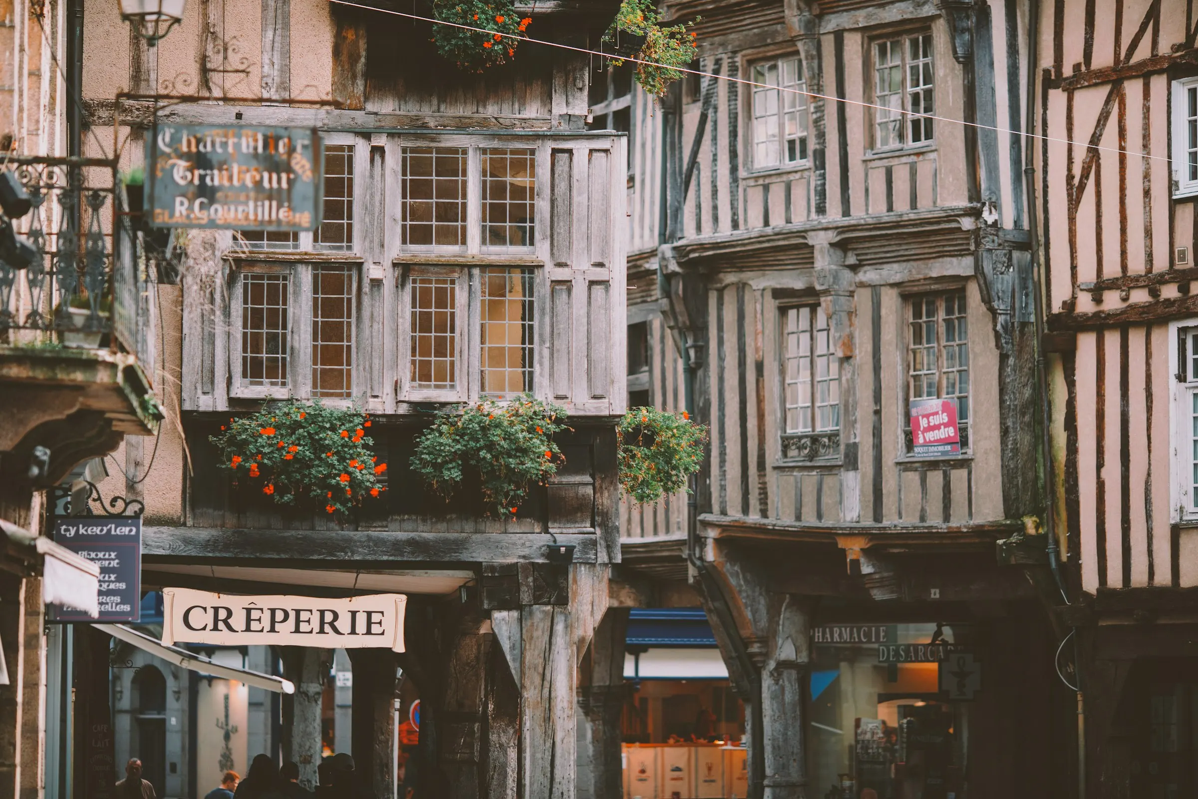 Close view of Dinan's overhanging medieval half-timbered facades with weathered timber, leaded windows, bright red geranium window boxes and old painted shop signs