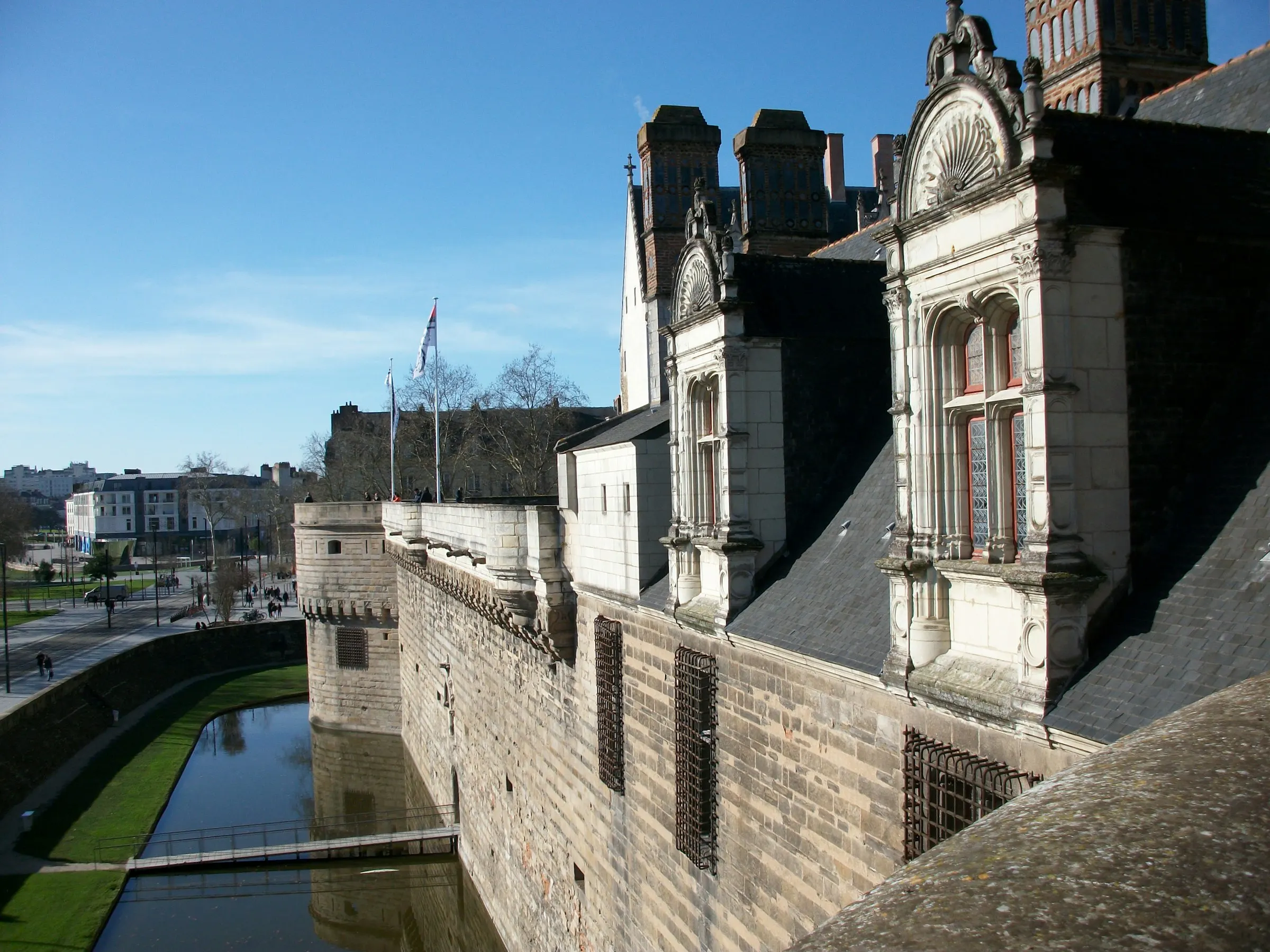 The moated Chateau des ducs de Bretagne in Nantes, its pale stone ramparts and ornate carved Renaissance dormer windows reflected in the water-filled moat under a clear blue sky.