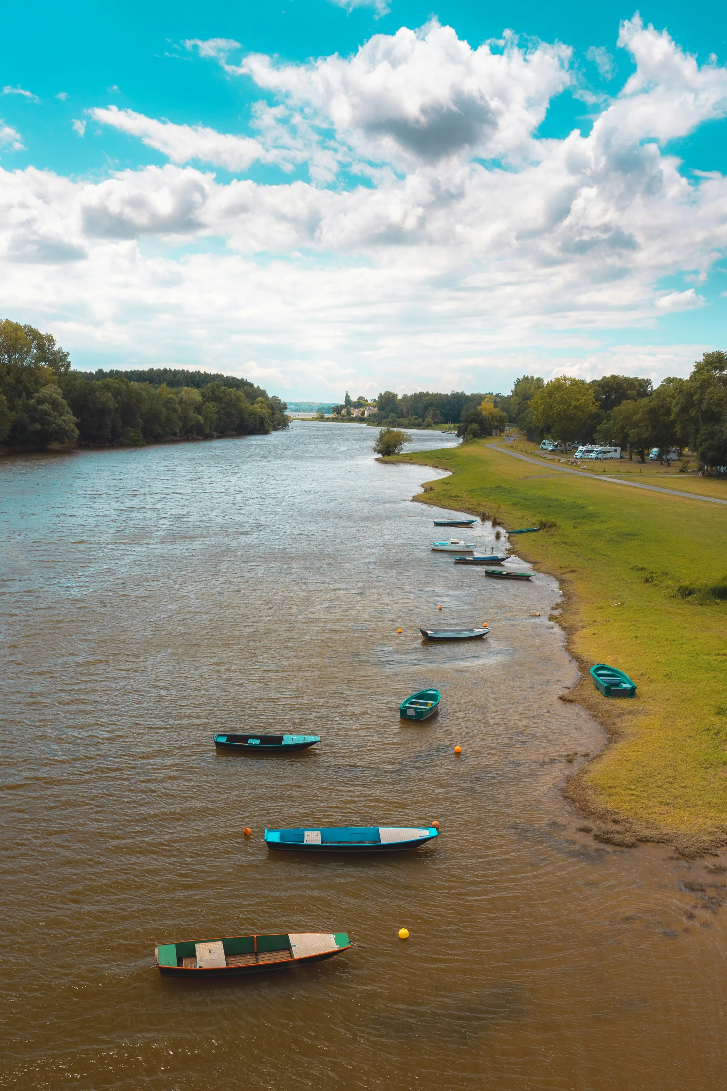 Traditional flat-bottomed wooden boats resting on the sandy banks of the Loire near Angers, with grassy riverside meadows under a bright, cloud-scattered summer sky.