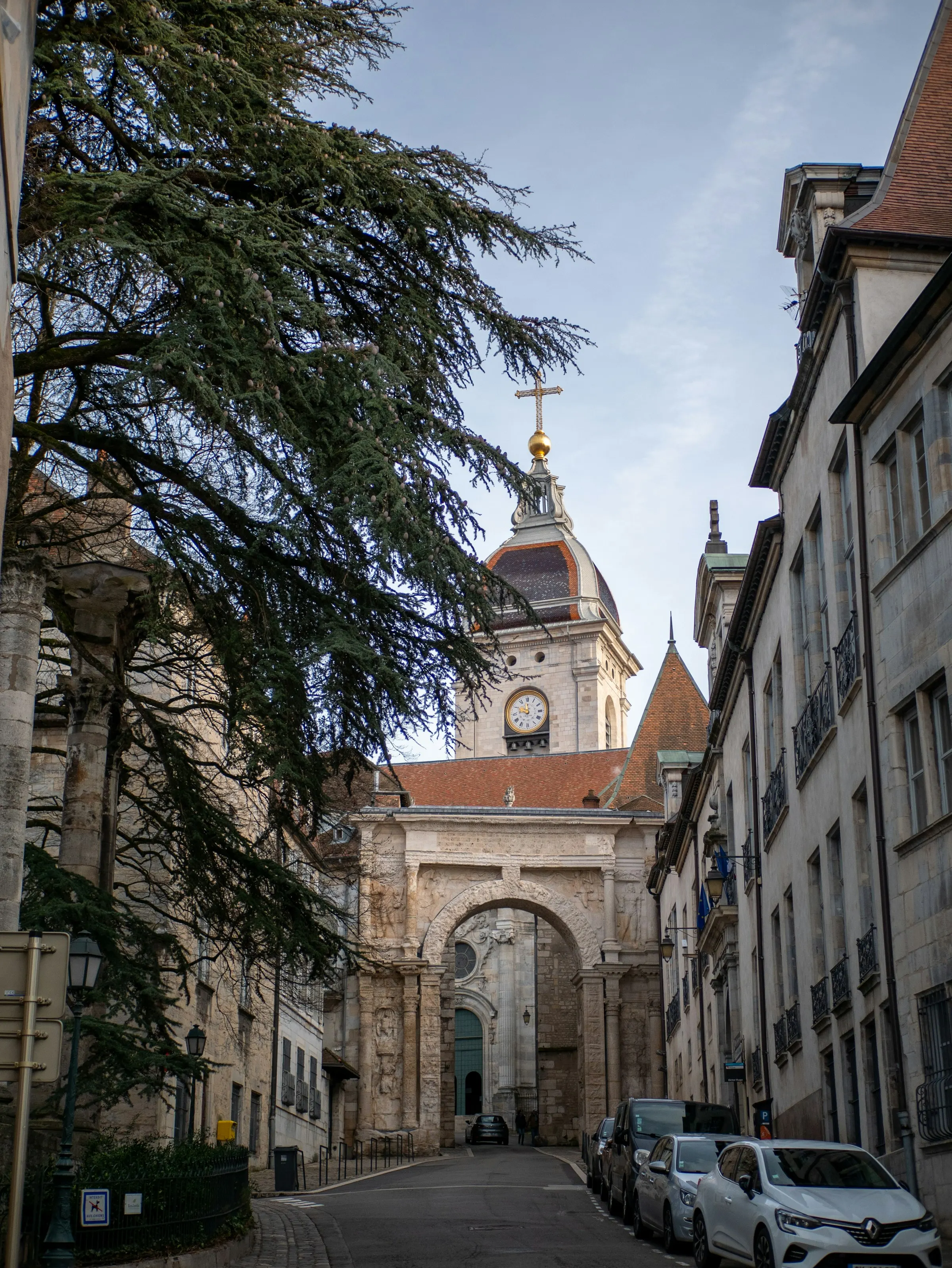 A quiet old-town street framed by a leaning cedar leads to Besançon's Roman Porte Noire arch and the domed, gold-cross clock tower of the Cathédrale Saint-Jean rising beyond.