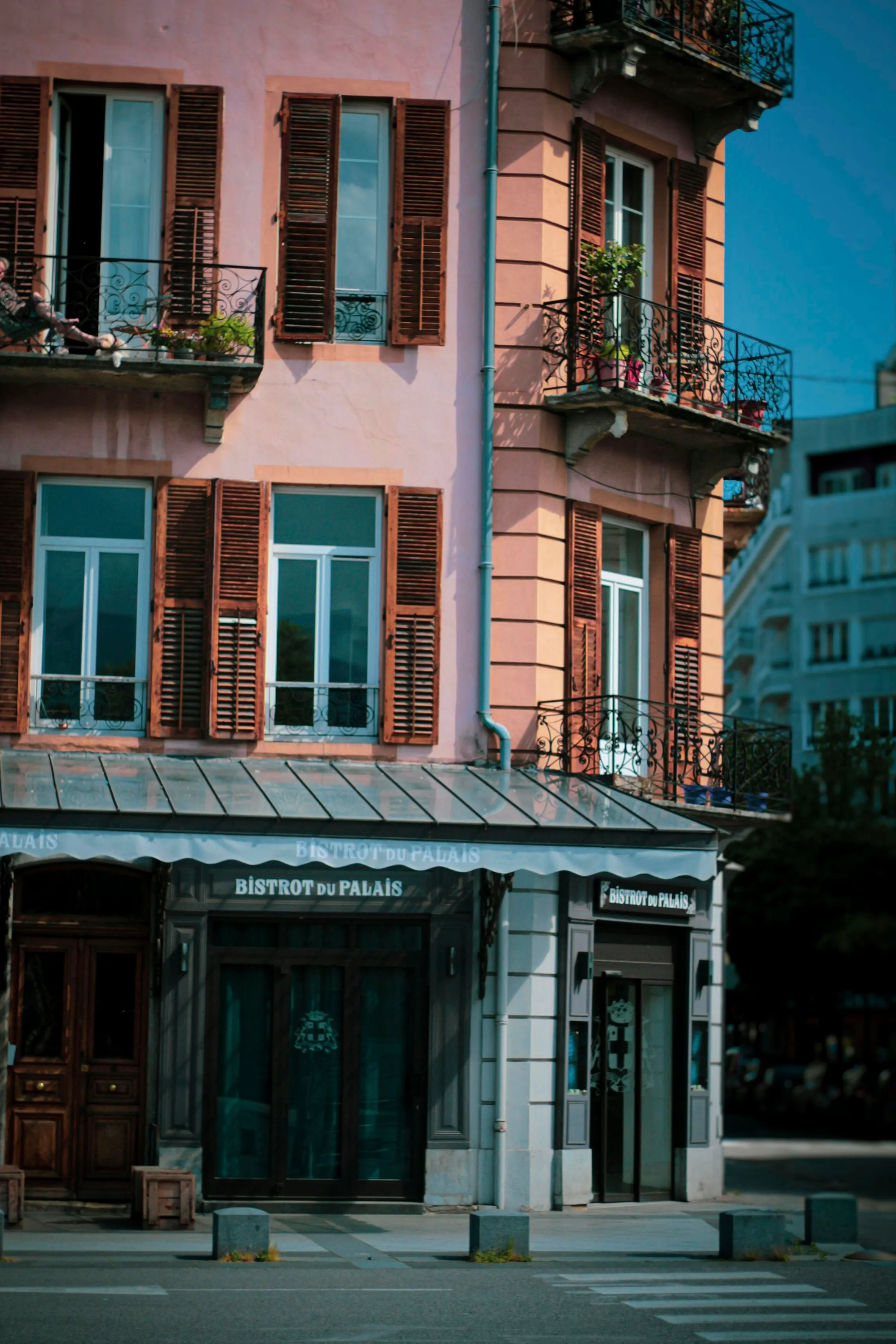 A sunlit salmon-pink corner building in Chambery housing the Bistrot du Palais, with brown louvered shutters and wrought-iron balconies of potted flowers under a deep blue sky.