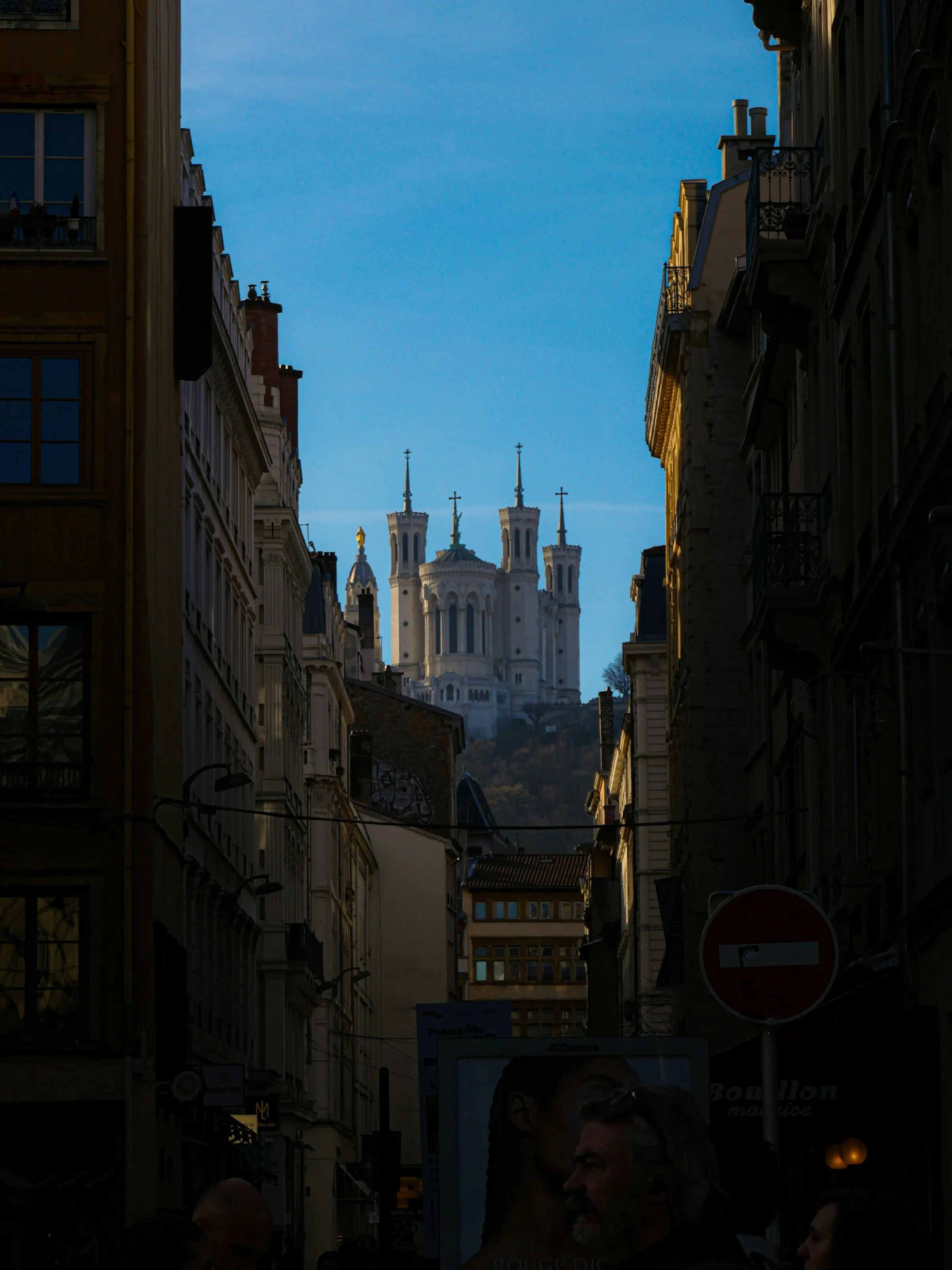 Looking up a narrow Vieux Lyon street in evening shadow toward the sunlit white towers of the Fourviere basilica on the hilltop, with passersby and a bouchon sign in the foreground