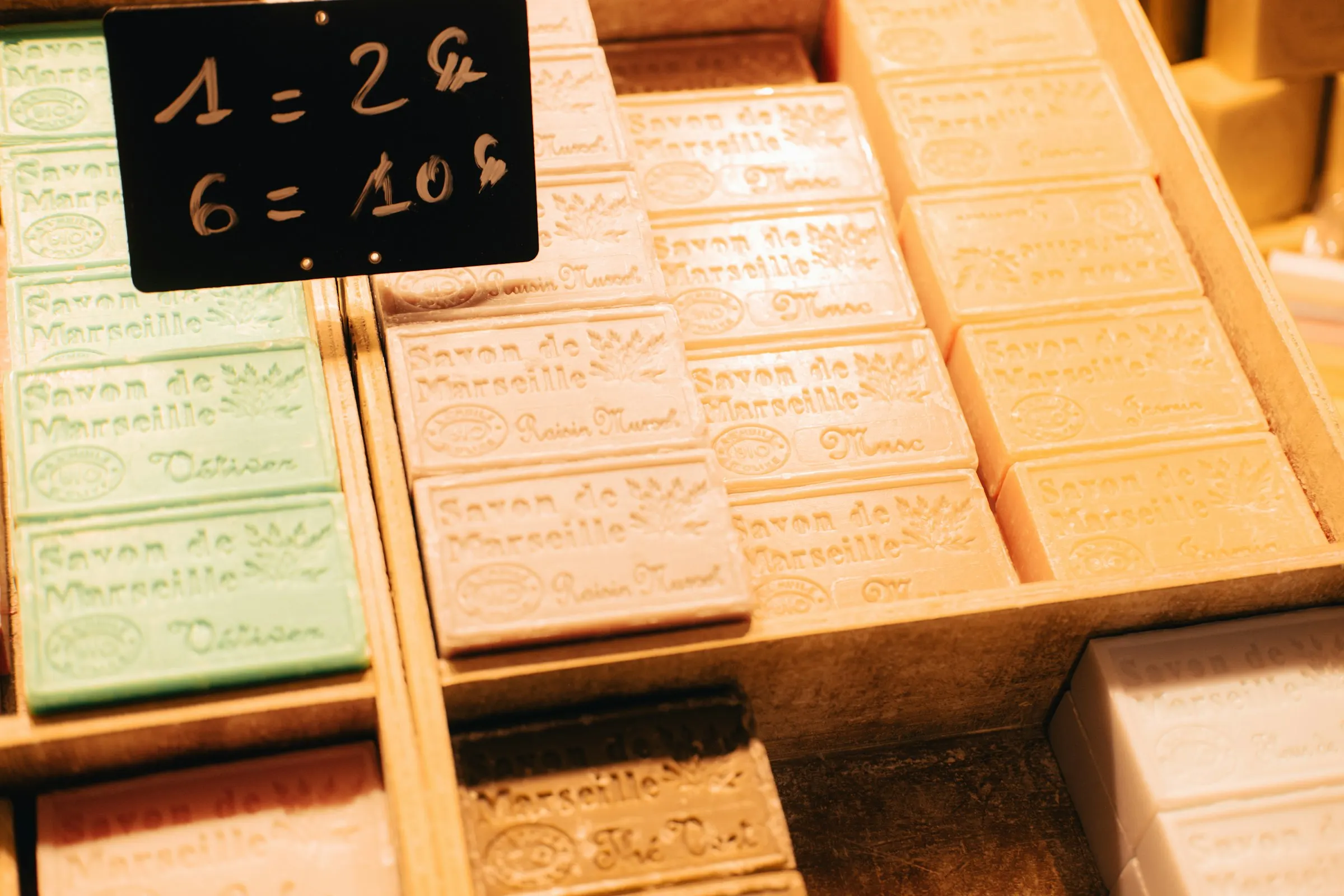 Rows of pastel green, cream and rose cubes of traditional Savon de Marseille soap, each block stamped with the town's name, stacked in wooden market crates beside a chalk price sign.