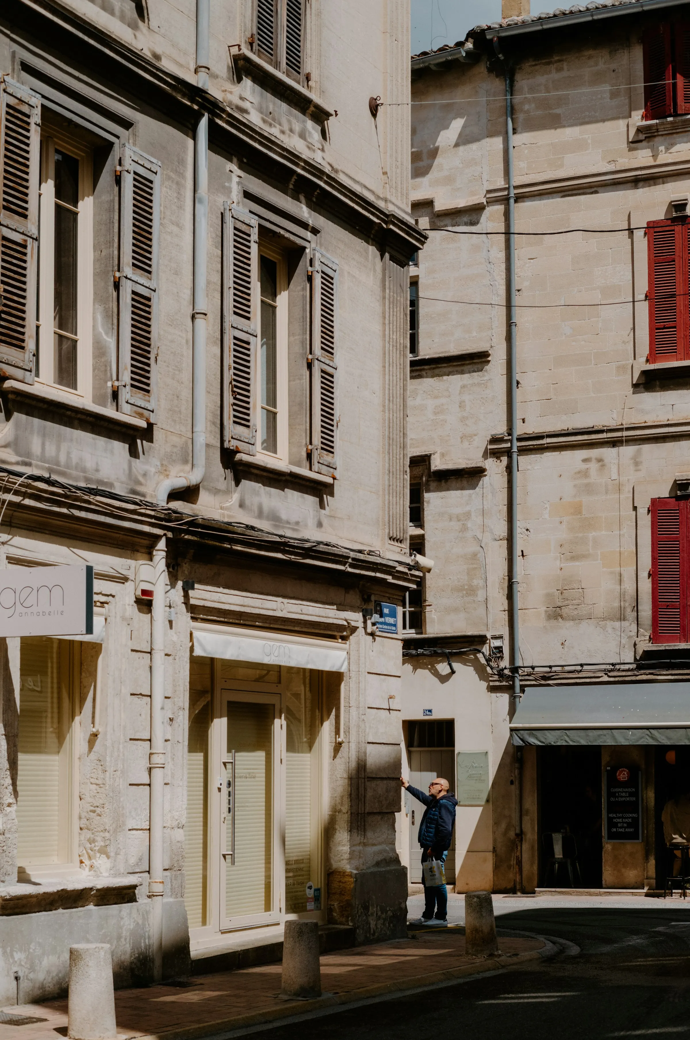 A sunlit corner of Avignon's old town on Rue Joseph Vernet: a man reaching up toward a wall plaque beside a pale boutique storefront, framed by tall honey-stone townhouses with weathered pale-blue and red shutters catching the afternoon light.