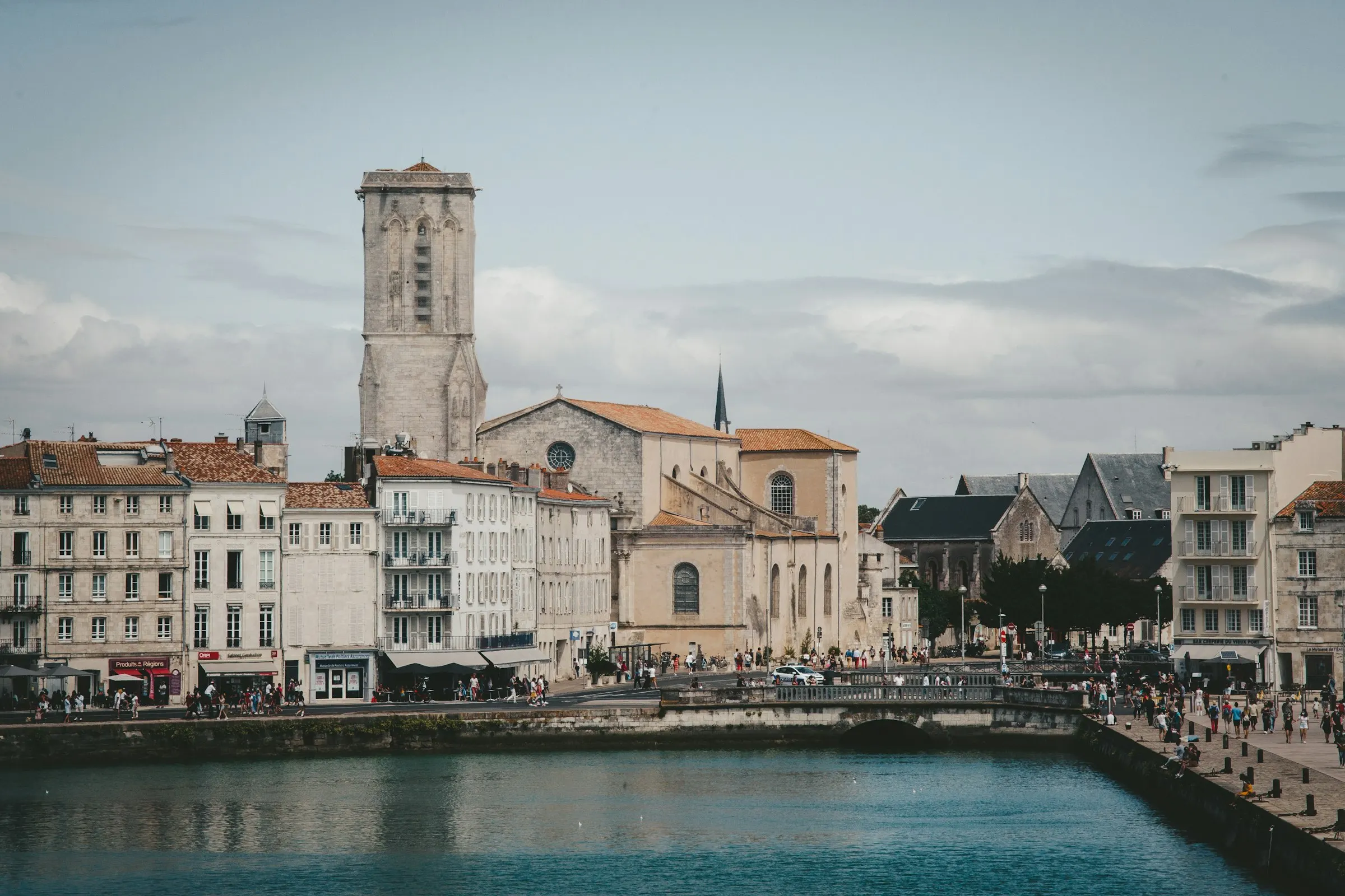 La Rochelle waterfront in soft daylight, the tall Gothic bell tower of the Eglise Saint-Sauveur rising above pale stone townhouses and a lively quay of shops and strollers beside the calm harbour.