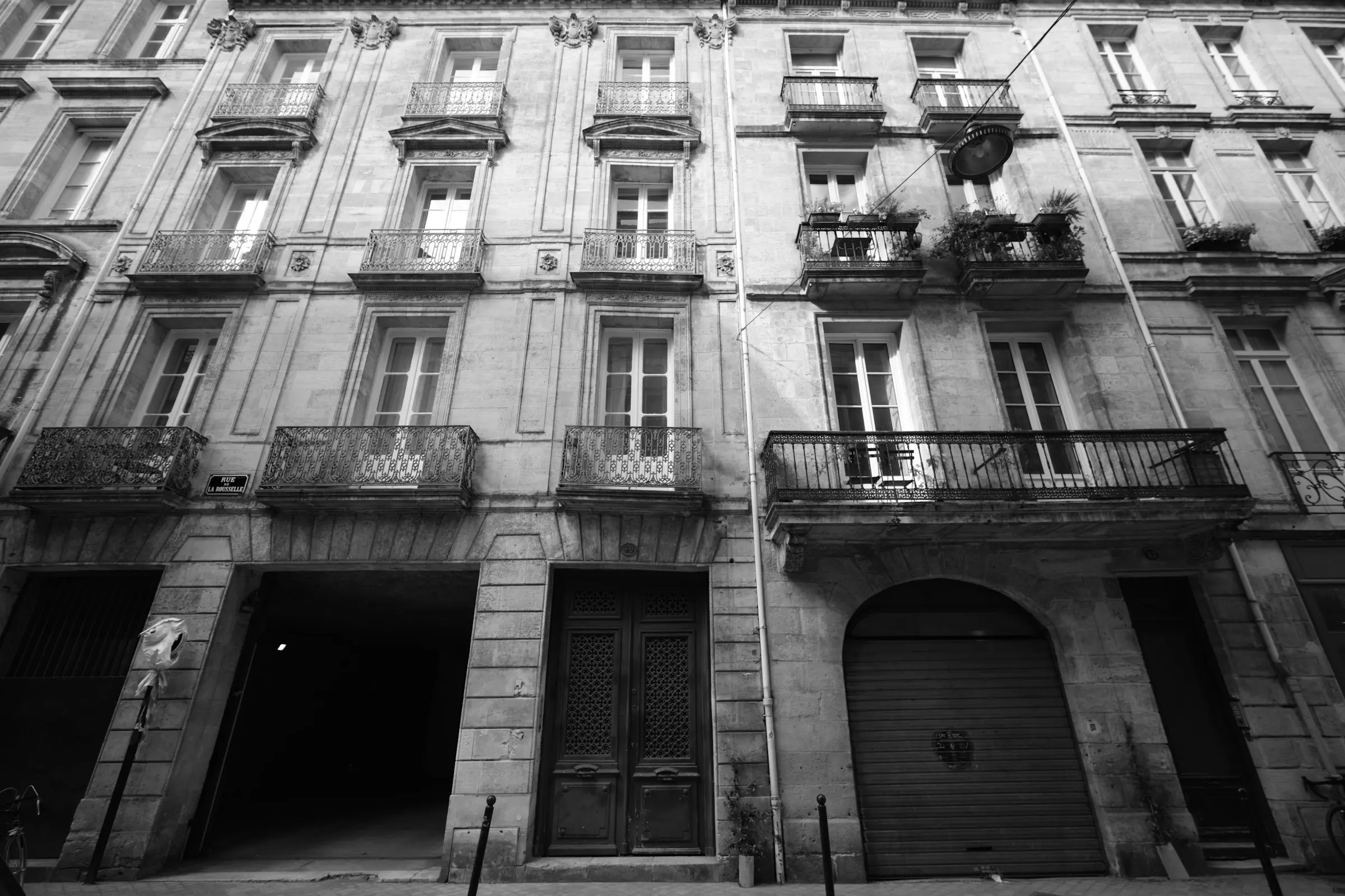 A low-angle black-and-white view of an elegant Bordeaux apartment building on Rue de la Rousselle, tiers of ornate wrought-iron balconies fronting tall shuttered windows in pale ashlar stone.