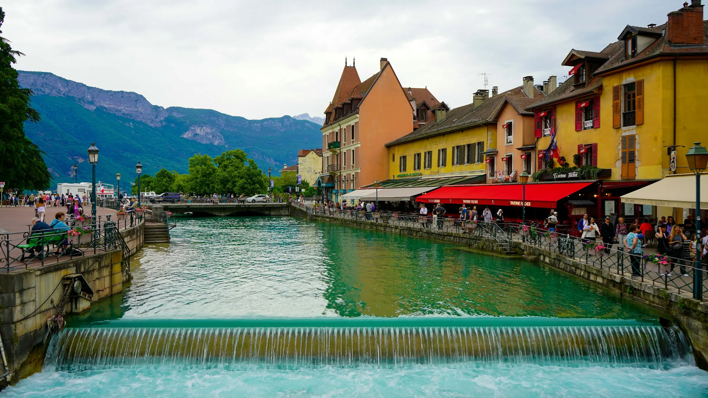 The Thiou canal weir in Annecy, colourful arcaded houses and cafe terraces with the mountains rising behind