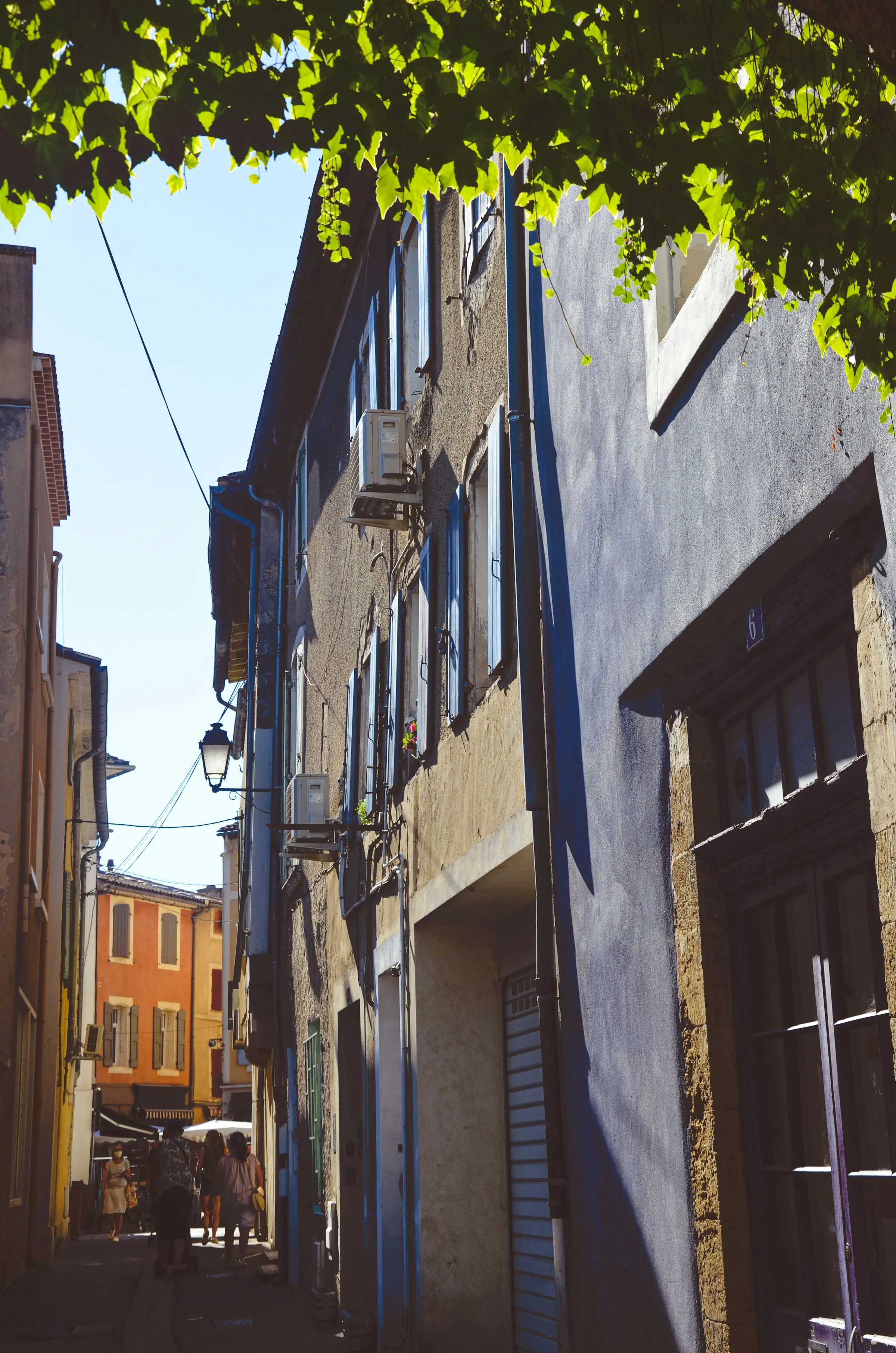 A sunny lane in a Provencal old town shaded by plane-tree leaves overhead, blue and grey shutters on stone houses and an ochre facade beyond, opening onto an open-air market with white umbrellas and shoppers at the far end.